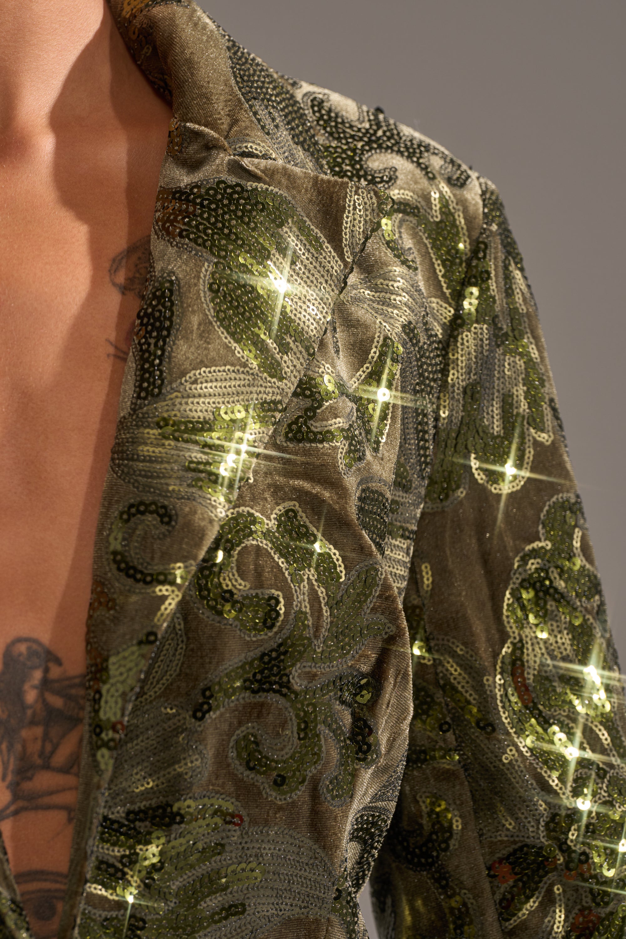 Close-up of a person wearing the AURORA SEQUIN VELVET BLAZER, featuring a deep V-neck and shimmering green sequins in an intricate pattern. A portion of their chest tattoo appears against a plain, neutral background.