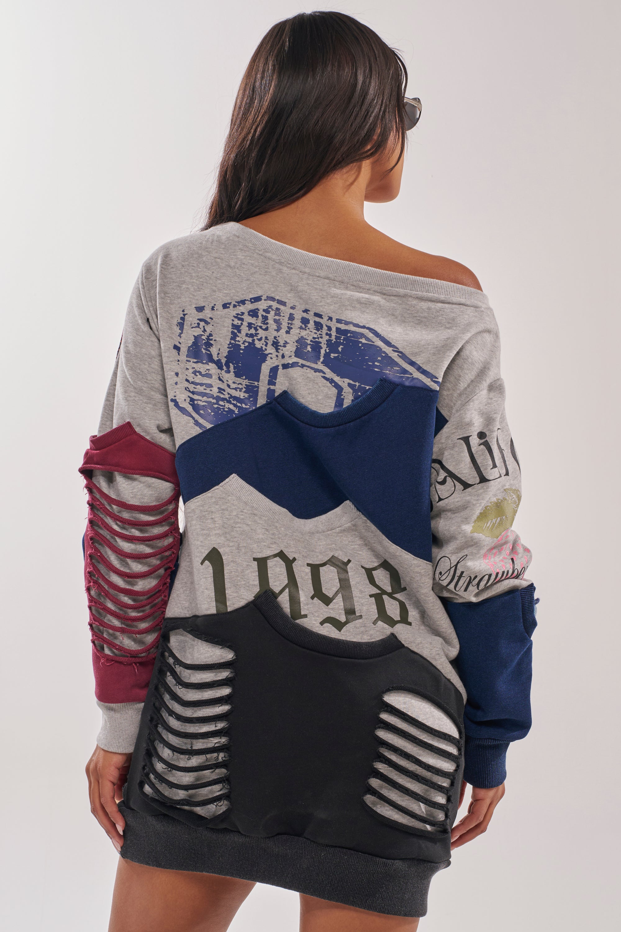 A woman with long dark hair stands with her back to the camera wearing the TALK LATER OFF THE SHOULDER PATCHWORK DRESS in gray, blue, and maroon, featuring graphic prints and slashed details on the sleeves and lower back. 4