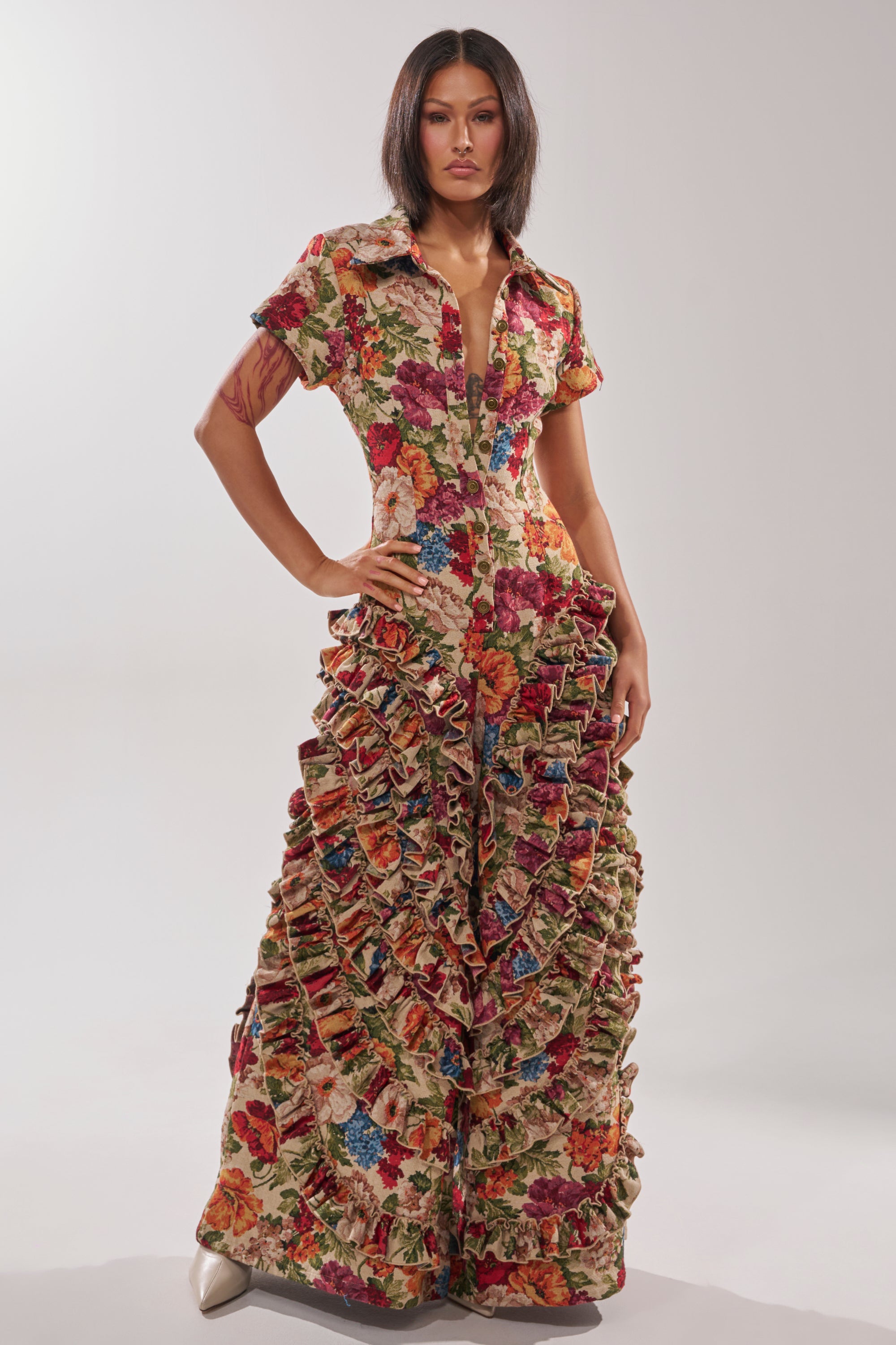 A woman poses confidently in the CRAZY IN LOVE FLORAL JUMPSUIT, featuring a vibrant, multicolored floral pattern, dramatic ruffle trim from the waist down, structured jacquard fabric, short sleeves, and a deep V-neckline.