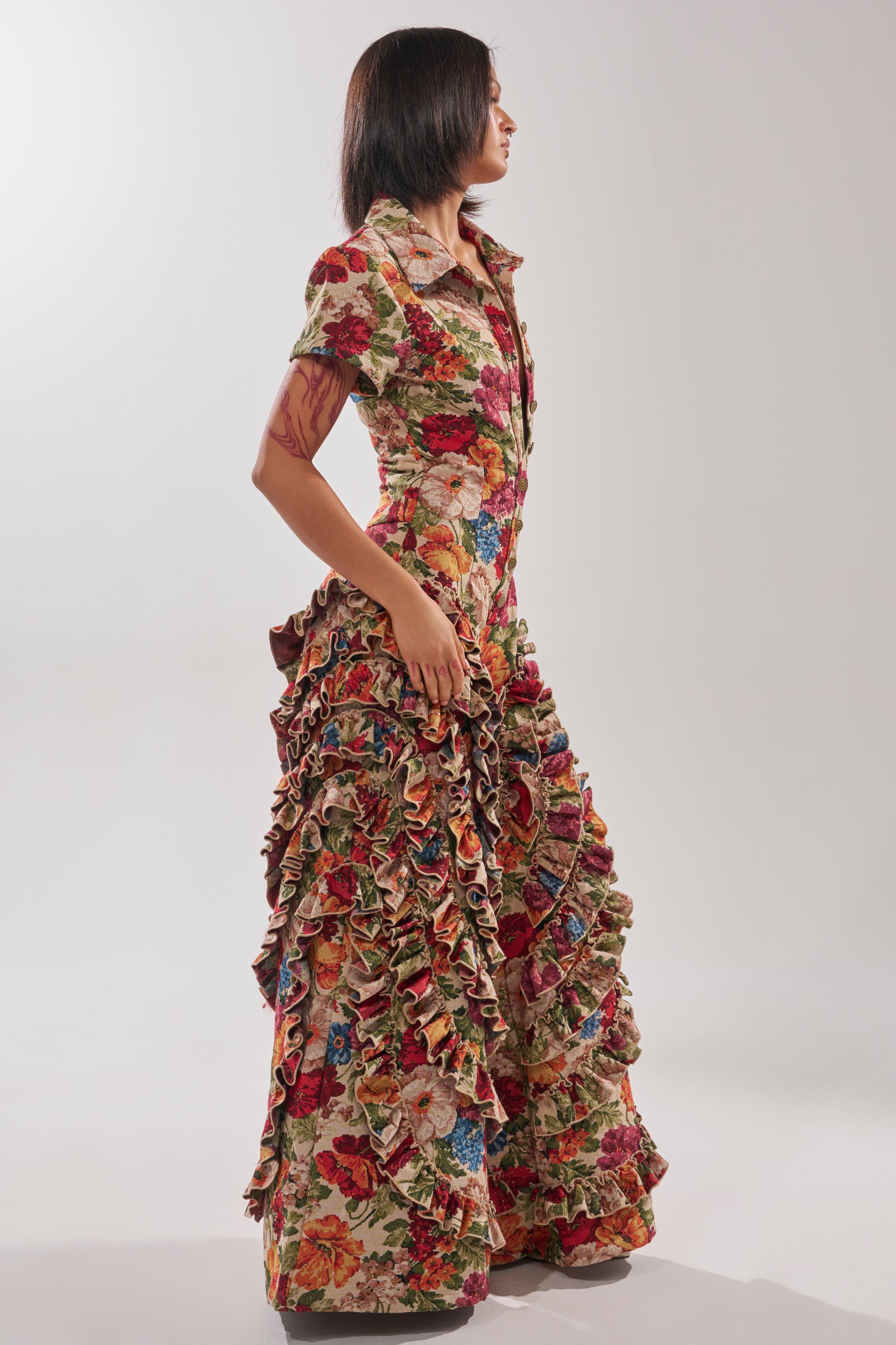A woman stands in profile wearing the CRAZY IN LOVE FLORAL JUMPSUIT, featuring a floor-length silhouette with floral print, short sleeves, and cascading ruffled trim, set against a plain, softly lit background.