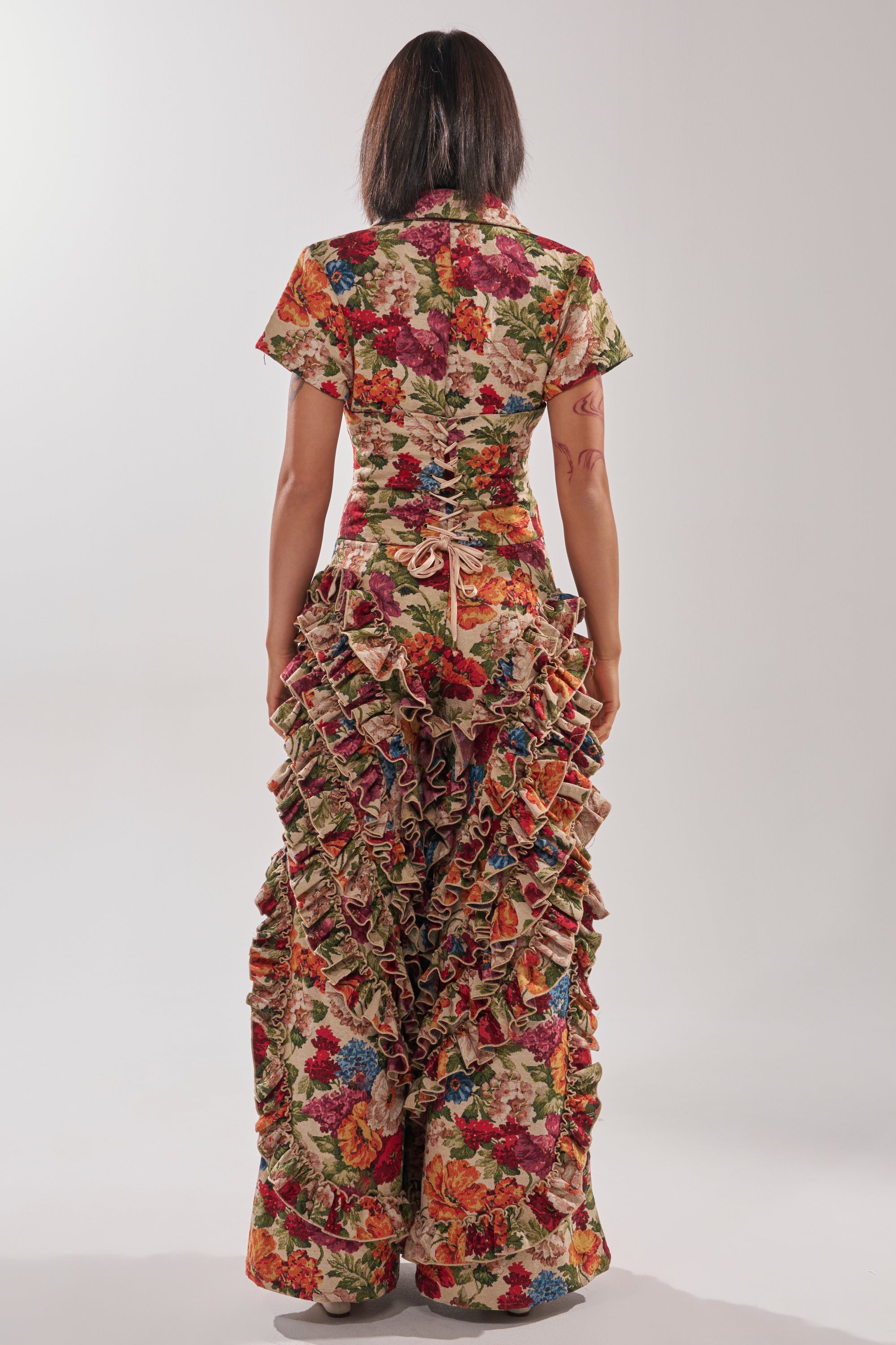 A person stands facing away, wearing the CRAZY IN LOVE FLORAL JUMPSUIT with a colorful floral pattern and dramatic ruffle trim. The jumpsuit features short sleeves and a fitted bodice, set against a plain, light background.