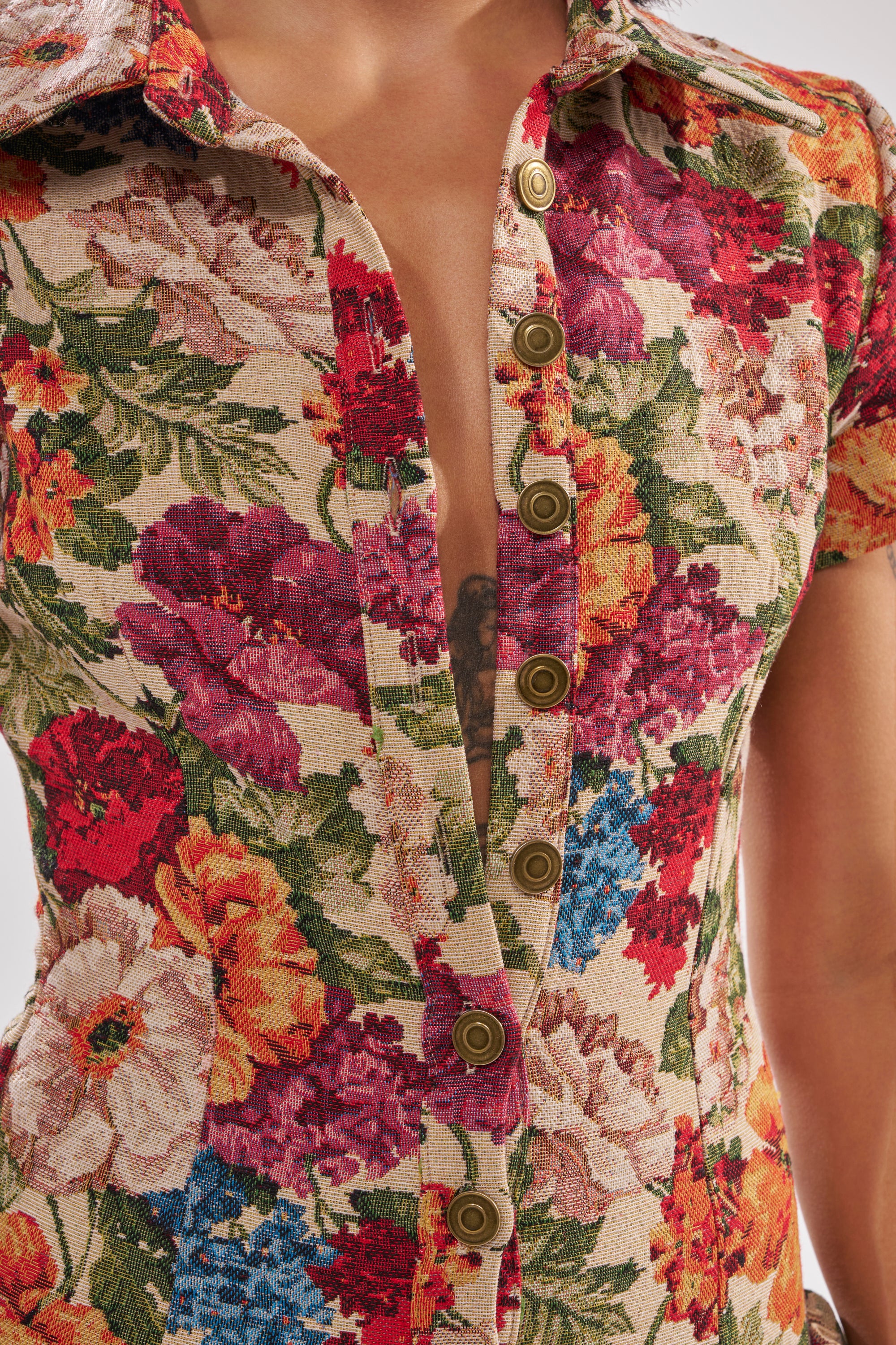 Close-up of a person wearing the CRAZY IN LOVE FLORAL JUMPSUIT, a beige jacquard one-piece with colorful floral patterns, front buttons, and a partially open neckline revealing part of a chest tattoo.