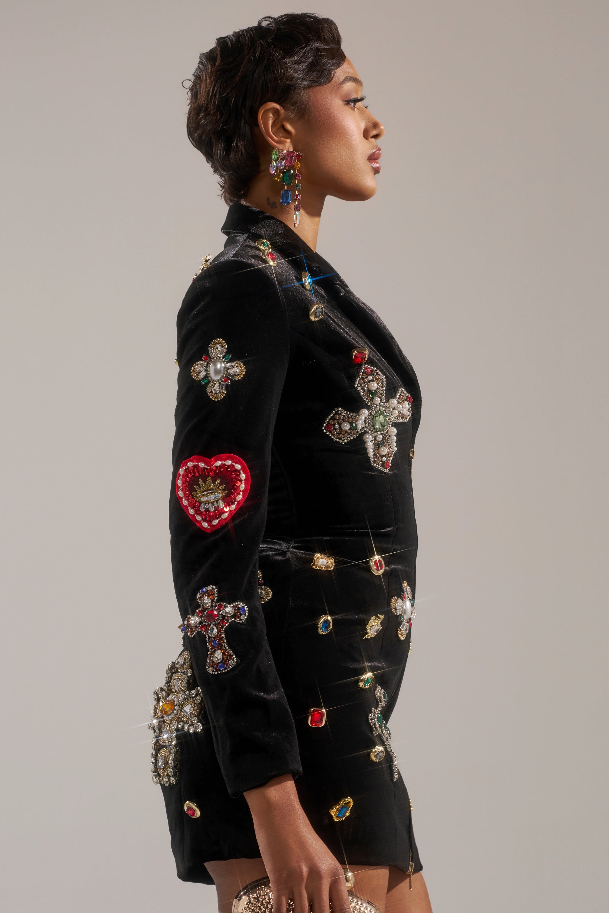 A person in profile wears the BUSINESS ONLY BLAZER DRESS, a black velvet piece decorated with jeweled crosses and hearts, paired with bold earrings, set against a plain background. 2