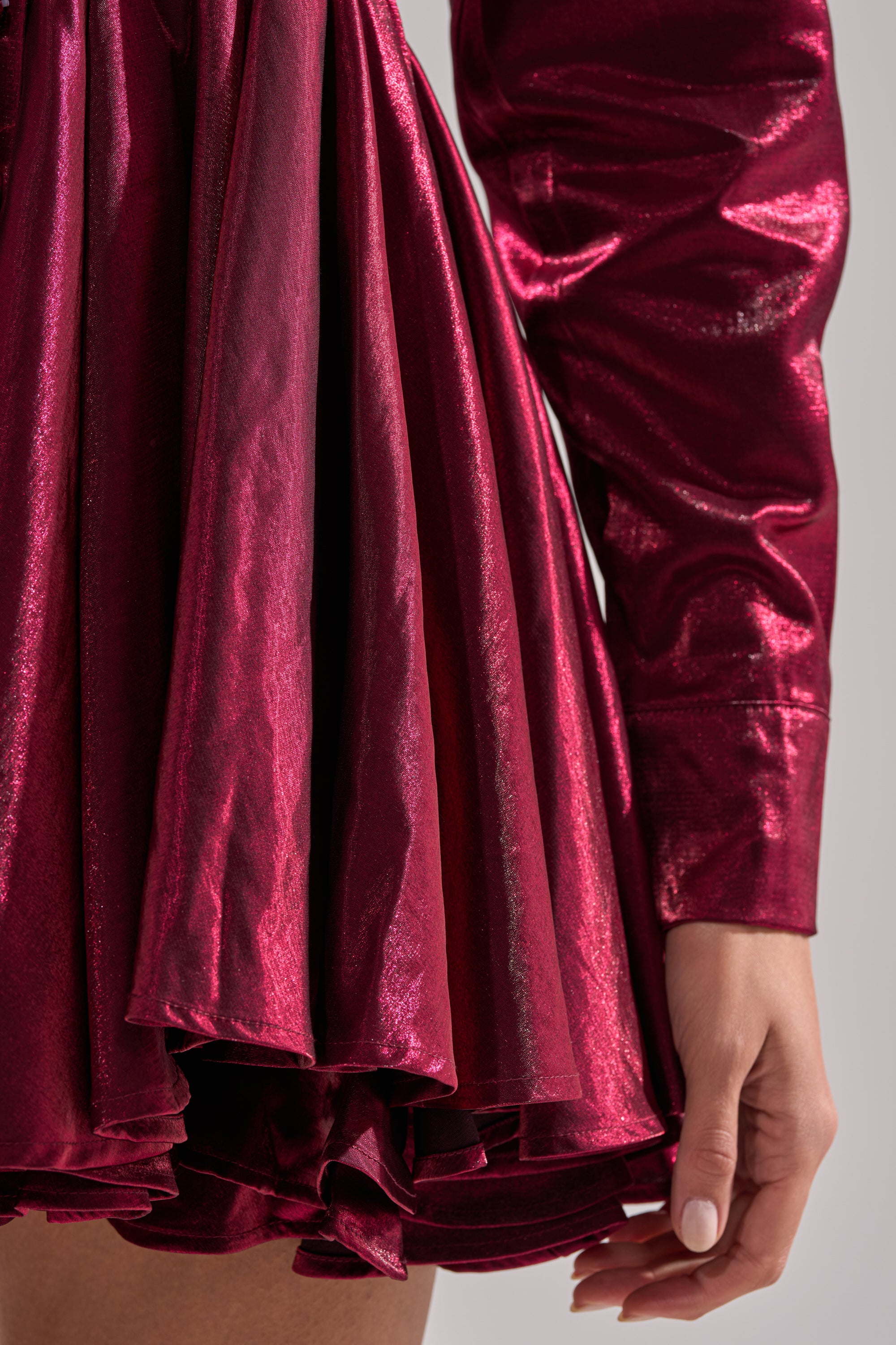 A close-up of a person wearing the OH MY BUTTON DOWN MINI DRESS—a shiny, deep red mini dress with long sleeves and a collar. Its glossy, textured fabric and ruffled, pleated skirt add dimension. The person's hand rests relaxed at their side.