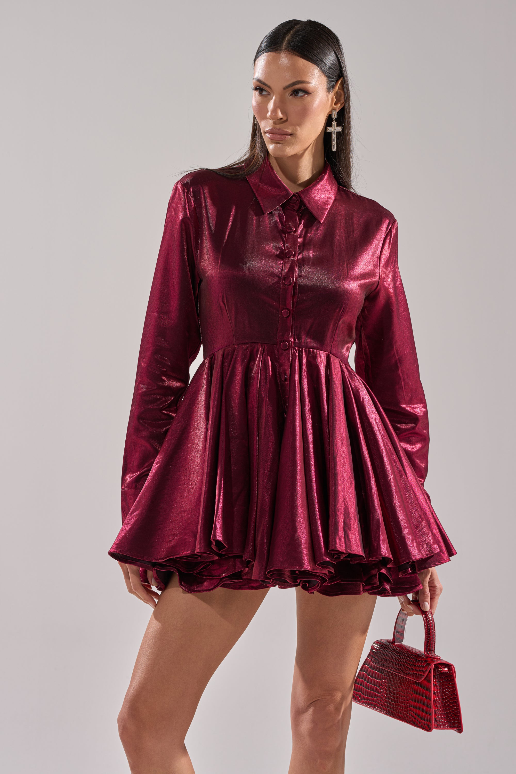 A woman wears the OH MY BUTTON DOWN MINI DRESS, a shiny deep red satin mini with long sleeves and a flared skirt. She holds a matching handbag, has long side-parted dark hair, large cross earrings, and stands before a plain light gray background.