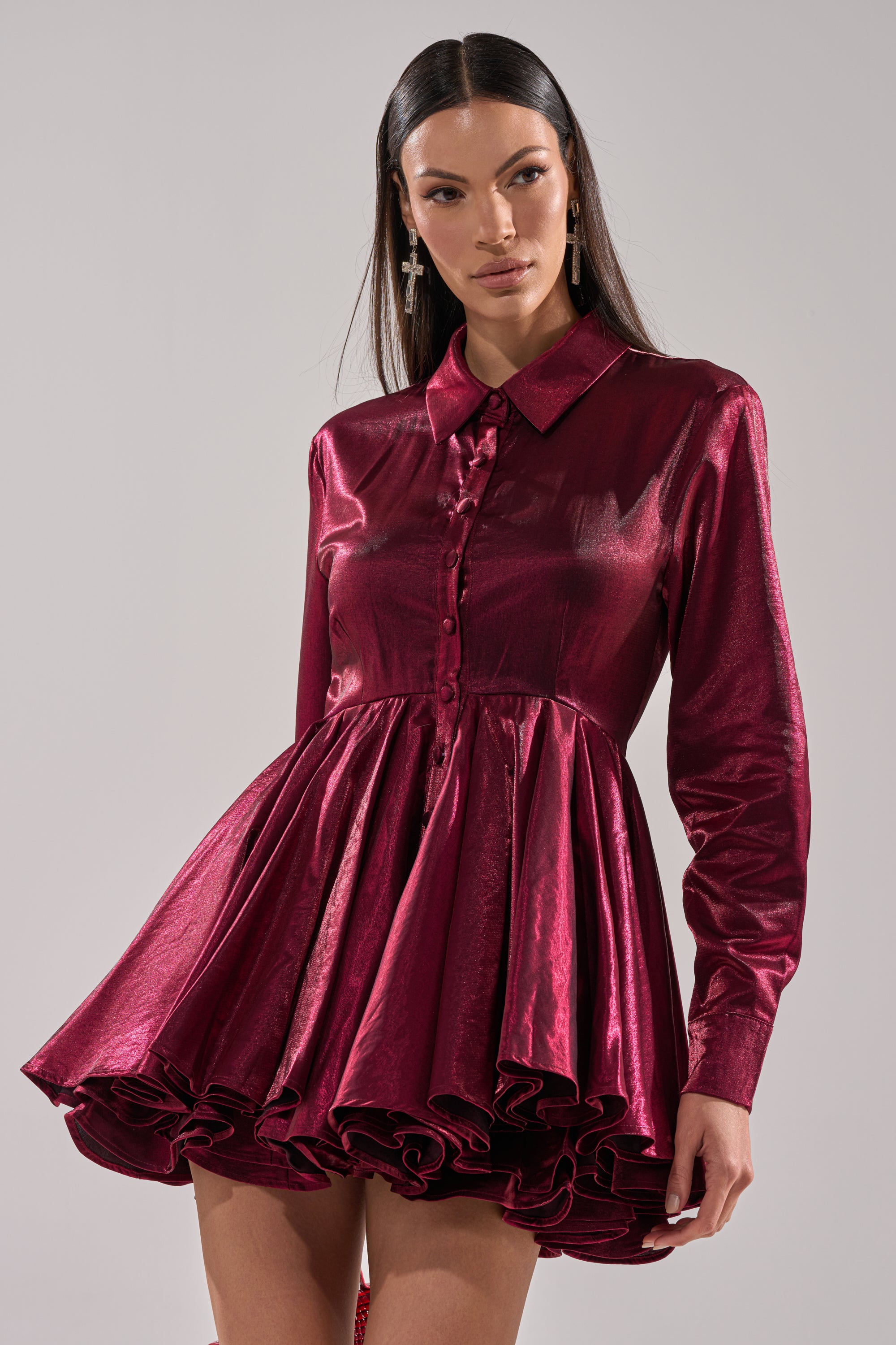 A woman wears the OH MY BUTTON DOWN MINI DRESS—a metallic deep red satin mini with long sleeves, button-down front, collared neckline, and a voluminous ruffled skirt. She has long straight hair and pairs it with striking cross earrings.