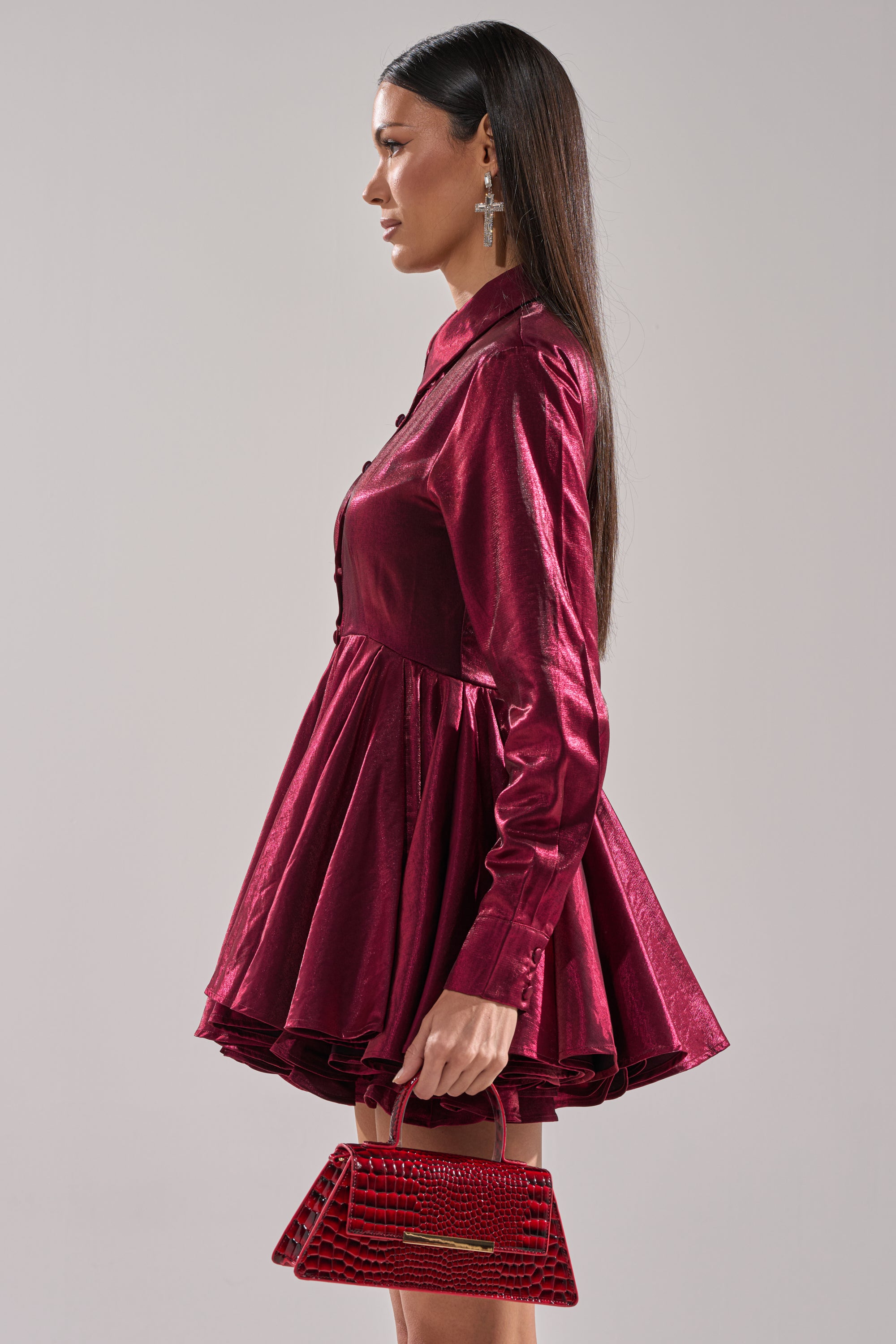 A woman in profile models the OH MY BUTTON DOWN MINI DRESS, a shiny deep magenta satin mini with long sleeves and a flared skirt. She holds a small red handbag, wears large cross earrings, and stands against a light gray background.