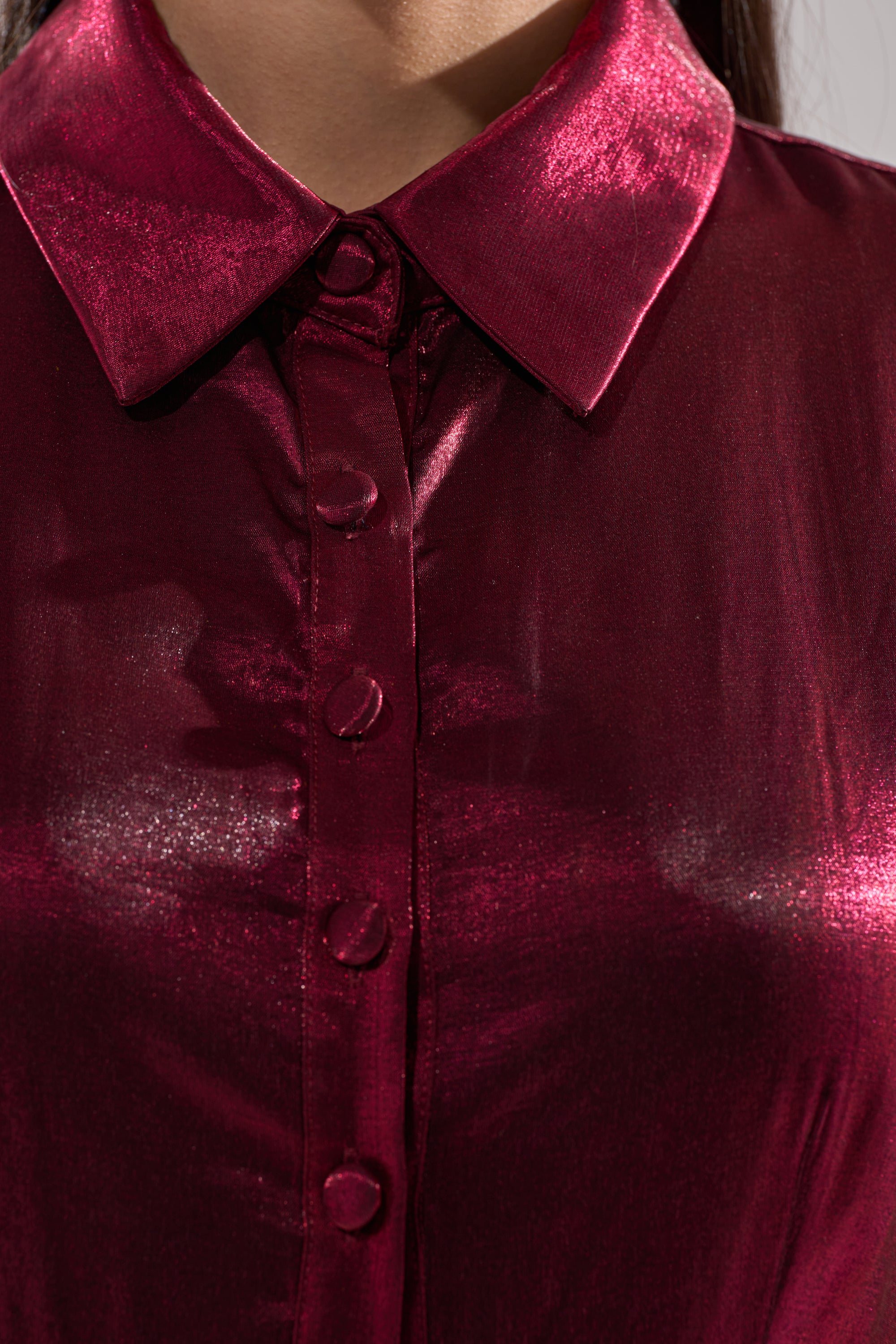 Close-up of a person wearing the OH MY BUTTON DOWN MINI DRESS in shiny metallic burgundy, featuring a pointed collar and glossy texture. Only the lower face and upper torso are shown.