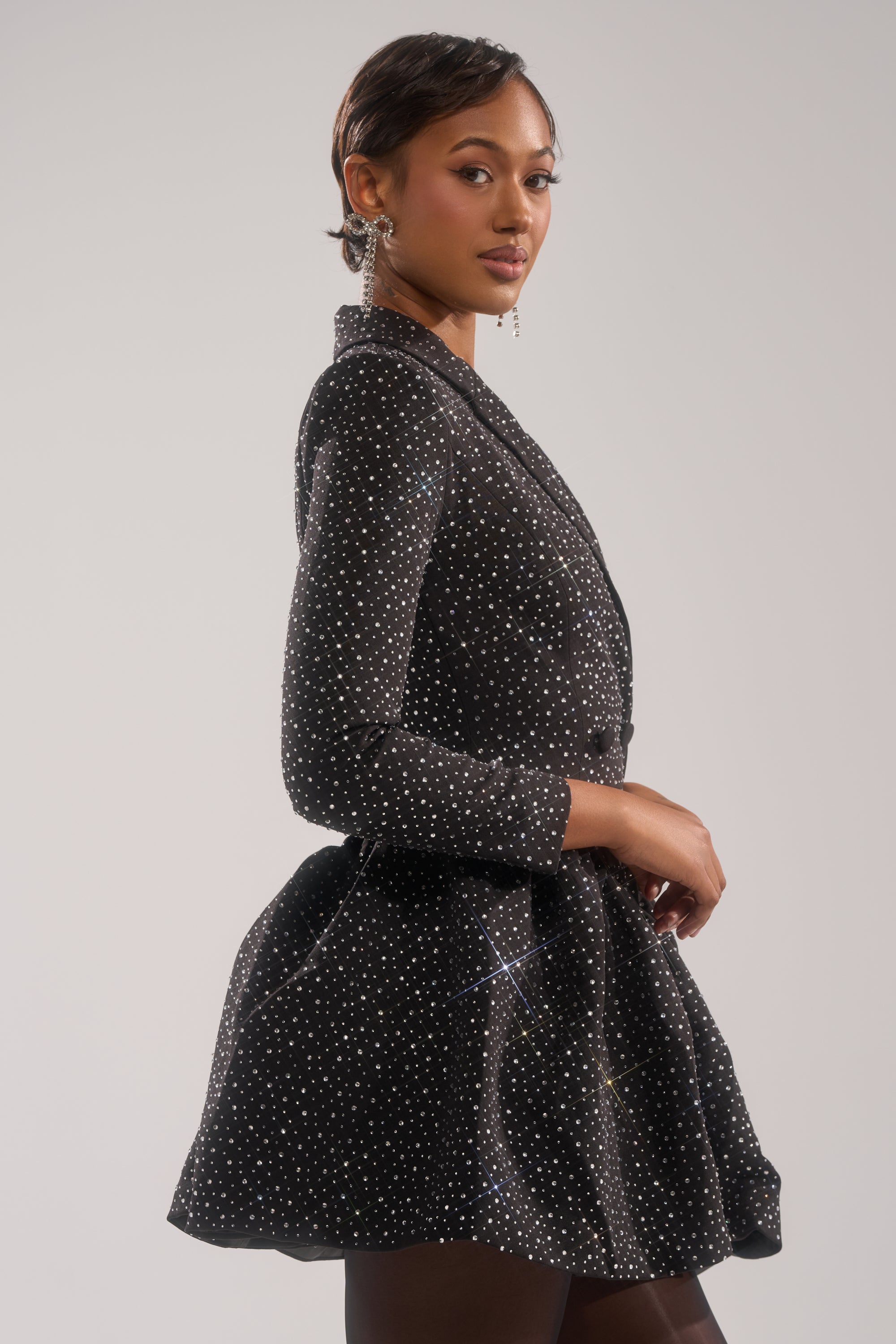 A woman with short hair and dangling earrings poses in the BACK IN BIZ MINI BLAZER DRESS by AKIRA Label—a sparkly black mini blazer dress featuring rhinestone embellishments and a flared skirt—against a plain, light background.