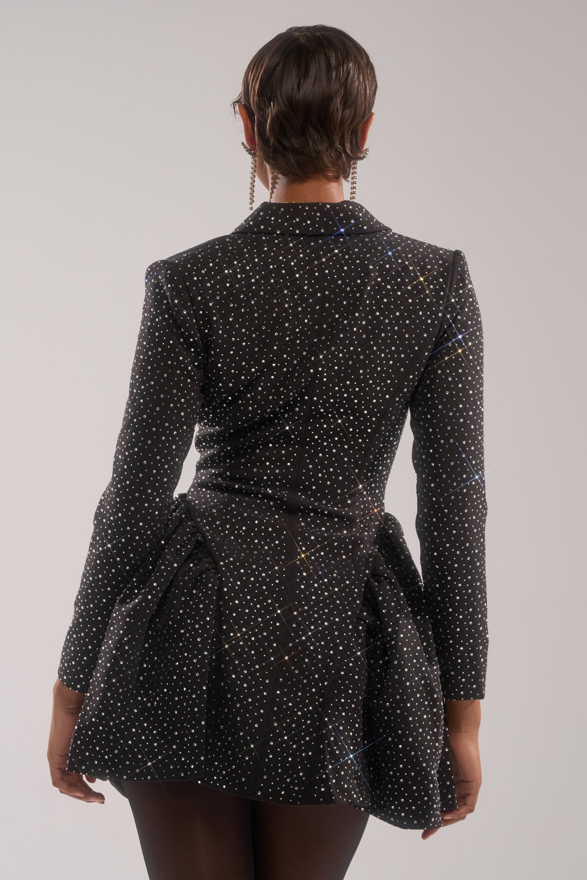 A person with short hair, seen from behind, wears the BACK IN BIZ MINI BLAZER DRESS—a fitted black mini blazer dress crafted from suiting fabric and adorned with sparkling rhinestone embellishments—against a plain light backdrop.