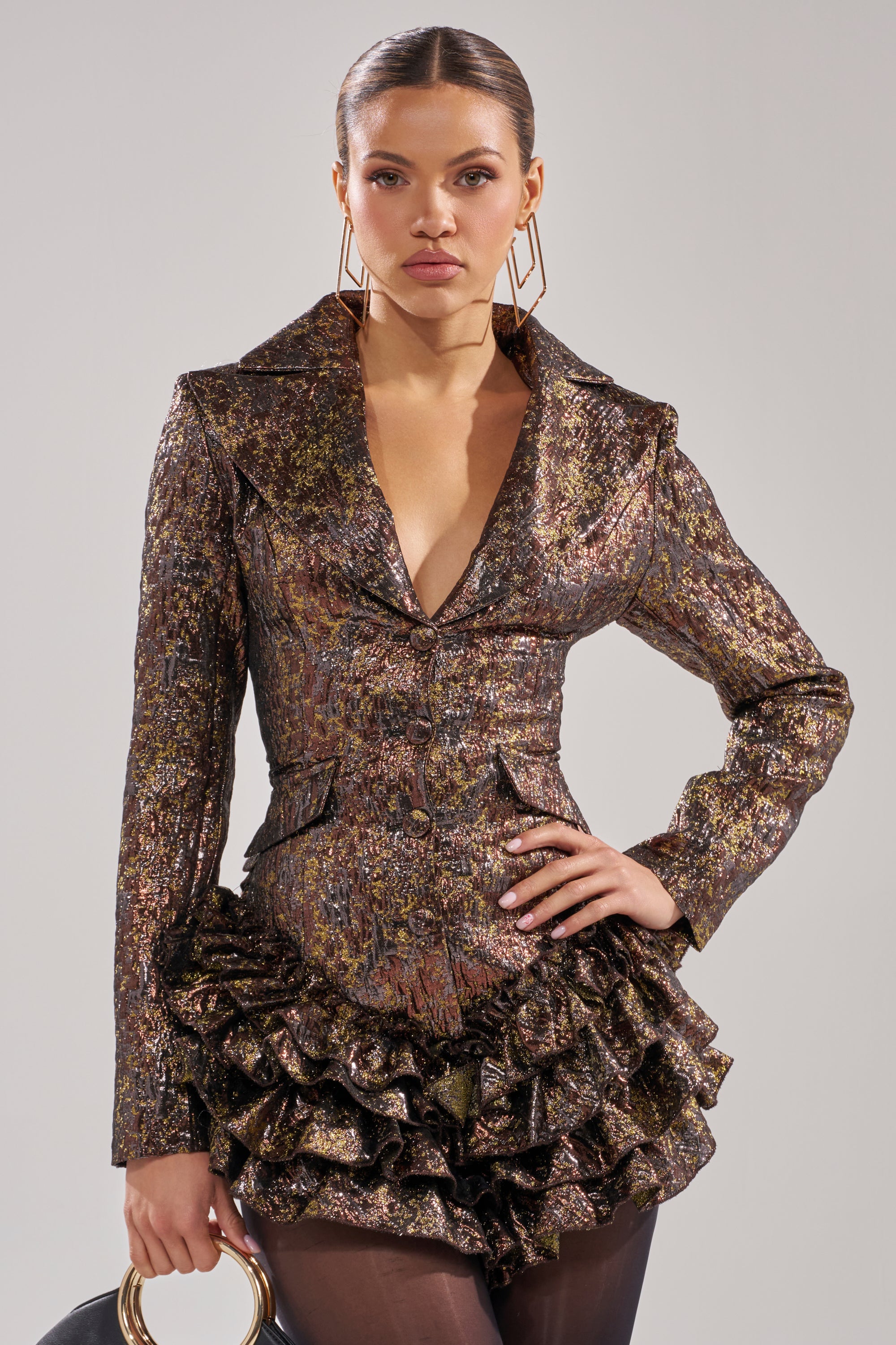 A woman wears the DANCING QUEEN BRONZE BLAZER ROMPER with structured shoulders and a ruffled skirt, styled with geometric earrings, a round-handled bag, and slicked-back hair for a bold, high-fashion statement.