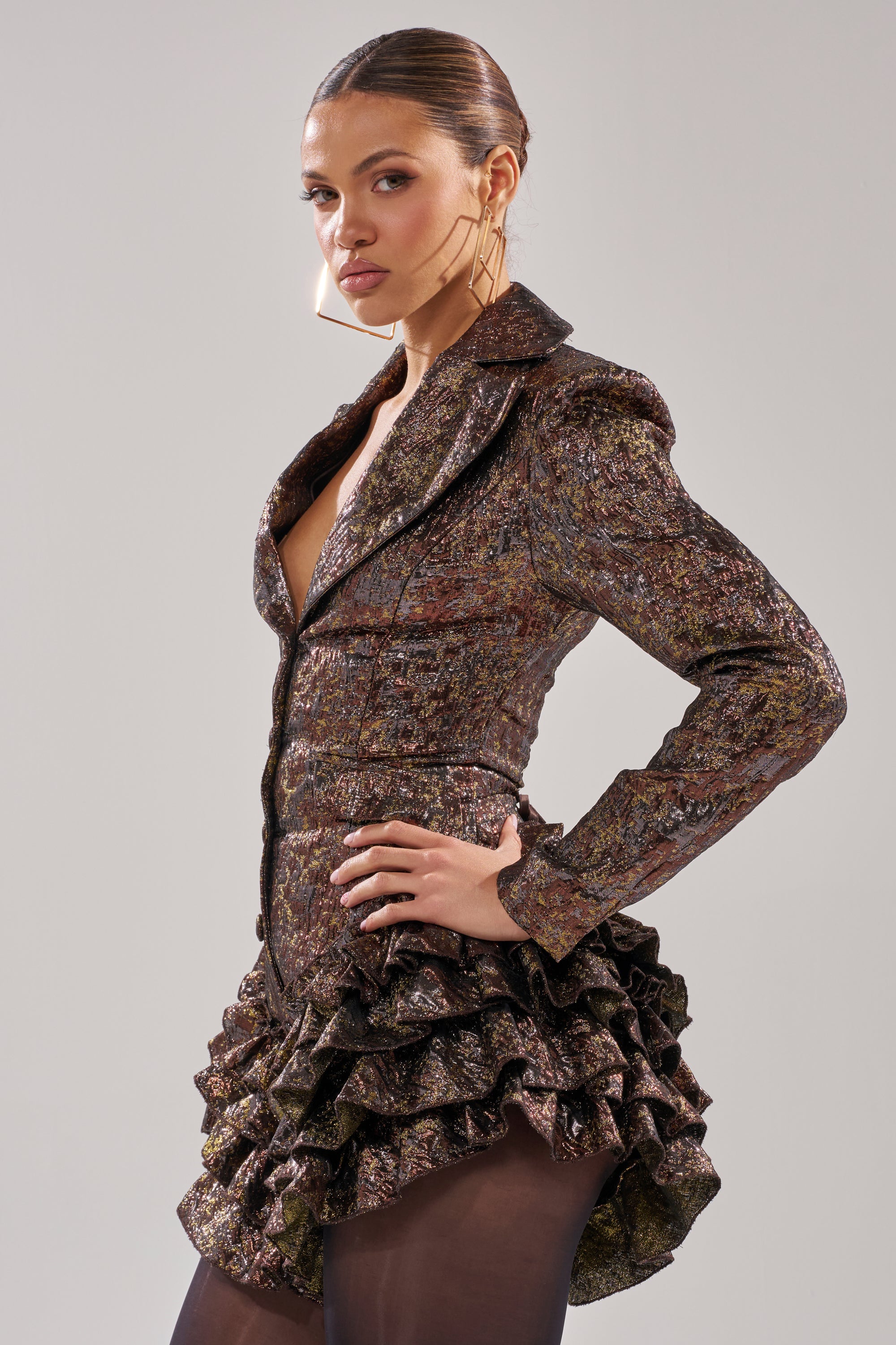 A woman models the DANCING QUEEN BRONZE BLAZER ROMPER with ruffled hem, sheer black tights, and large geometric earrings. Her sleek hair is pulled back as she poses against a plain gray background.