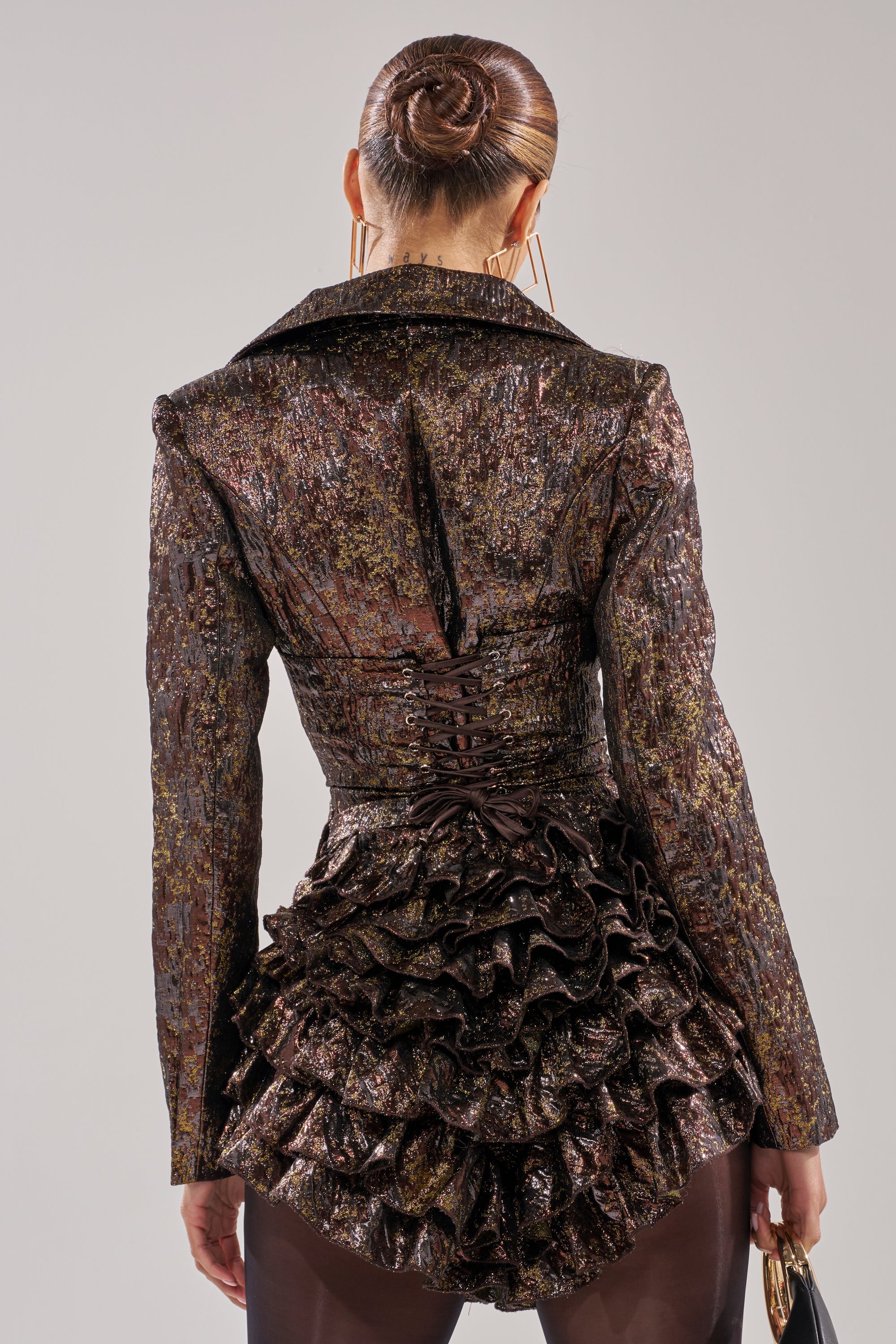 A person with slicked-back hair in a bun stands against a plain background, wearing the DANCING QUEEN BRONZE BLAZER ROMPER featuring a textured, metallic finish and structured shoulders.