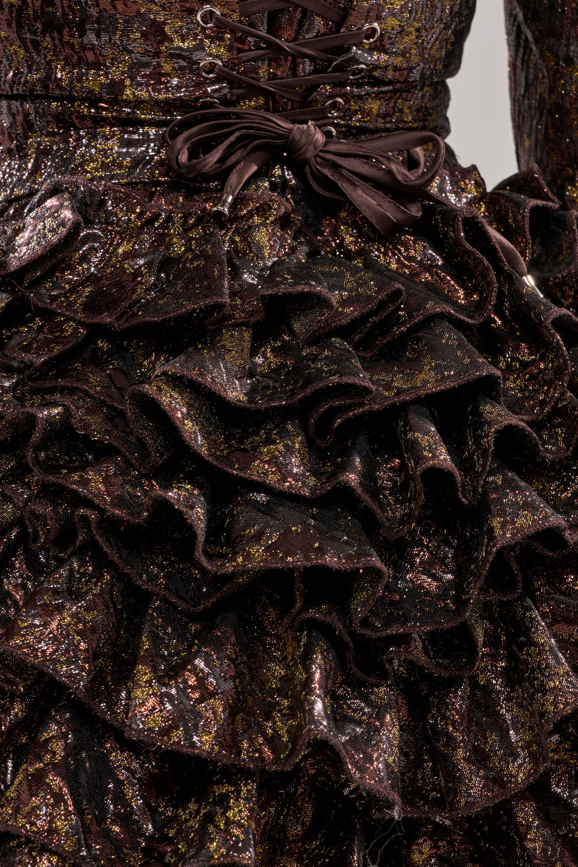 Close-up of the DANCING QUEEN BRONZE BLAZER ROMPER: features ruffled metallic fabric in dark brown and gold, a decorative bow, lace-up waist detail, and a shiny textured finish.