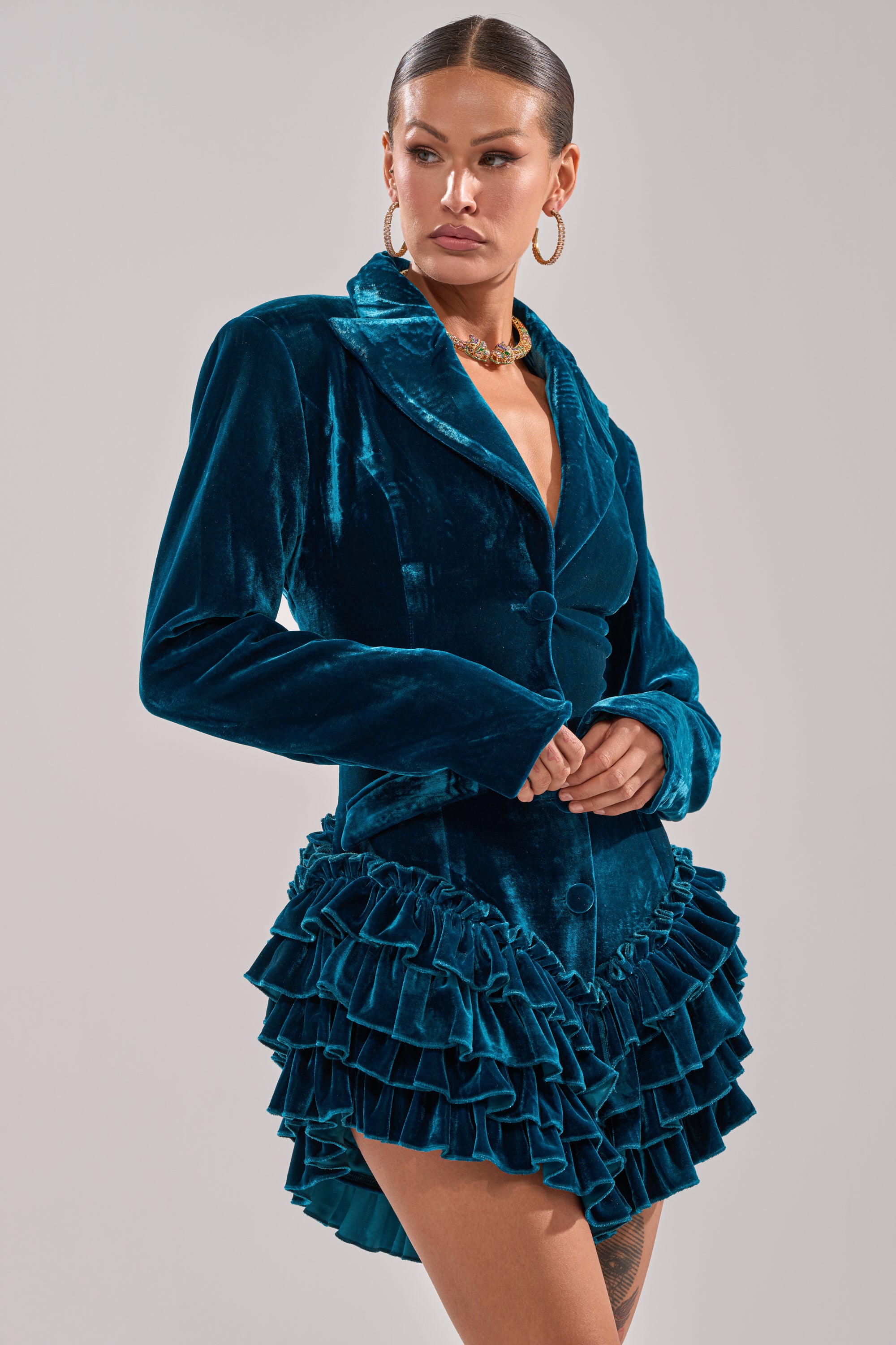 A woman models the SHE'S ROYALTY VELVET ROMPER in teal crushed velvet, featuring a blazer-style top with a deep V-neck and tiered ruffle mini skirt hem, standing against a plain light gray background.