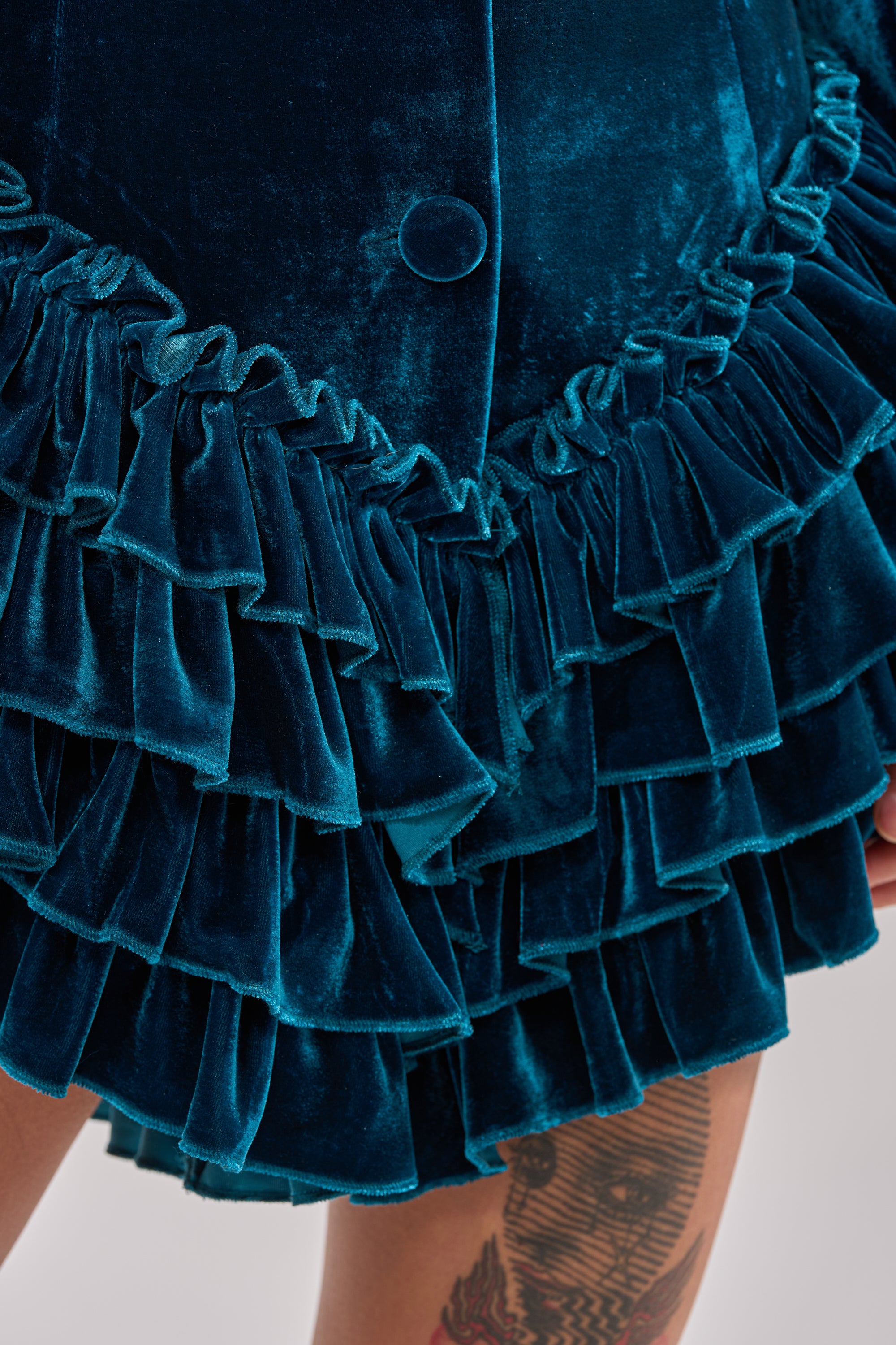 Close-up of the SHE'S ROYALTY VELVET ROMPER in teal crushed velvet, featuring a tiered ruffle hemline and button detail, with part of a tattooed leg visible.