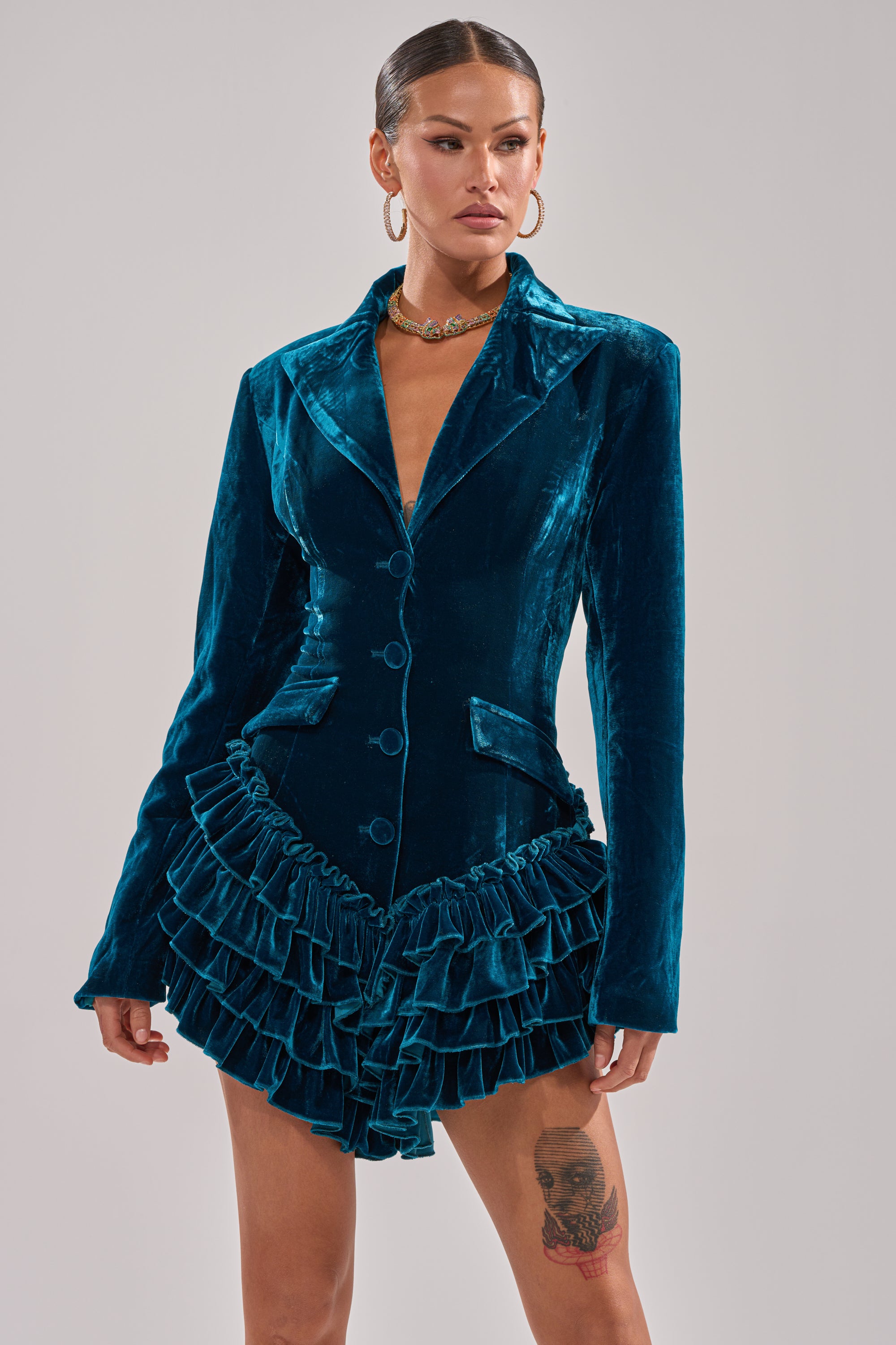 A woman stands confidently in the SHE'S ROYALTY VELVET ROMPER, featuring teal crushed velvet and a tiered ruffle hemline. She accessorizes with slicked-back hair, bold earrings, a gold necklace, and reveals a skull tattoo on her thigh.