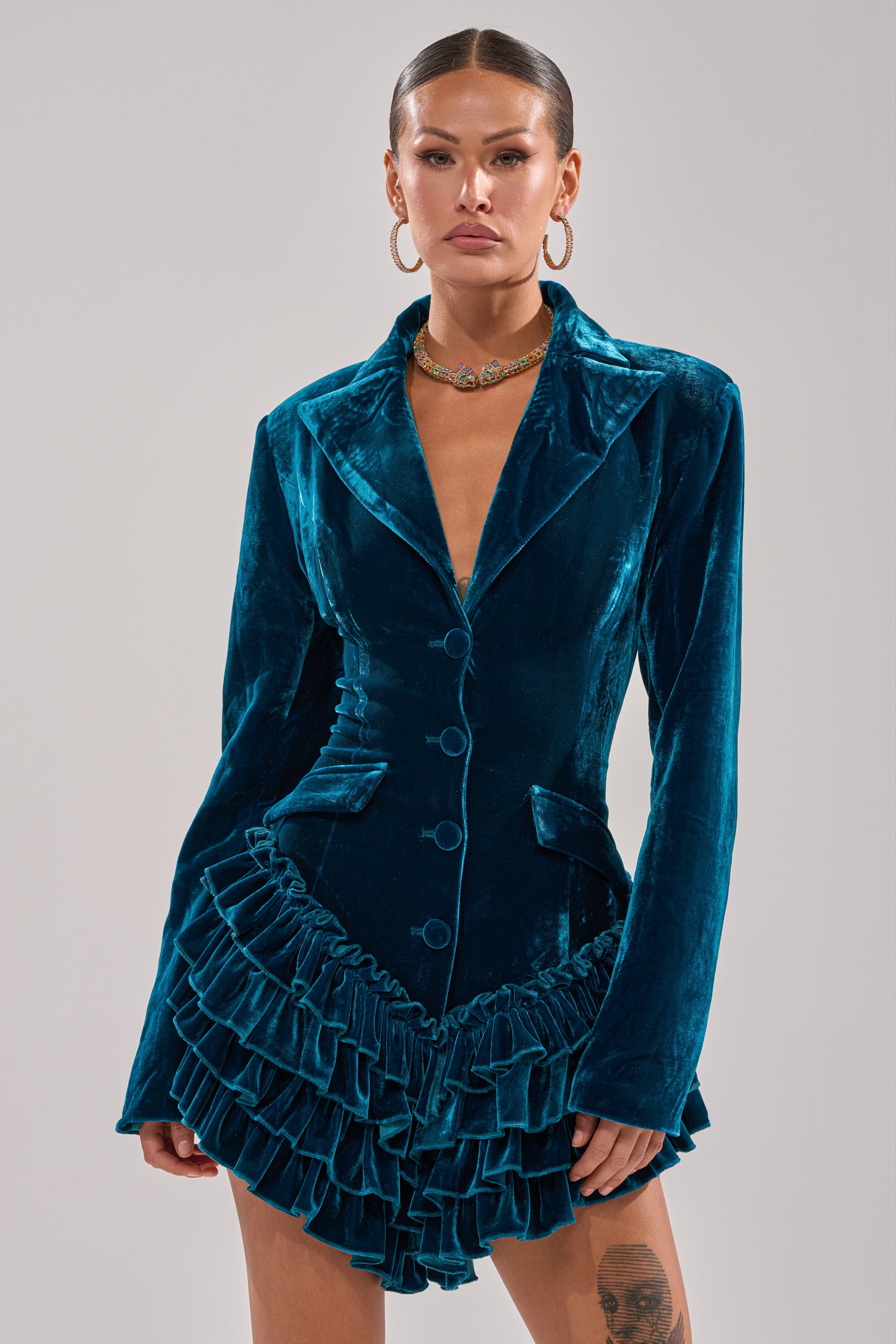 A woman in the SHE'S ROYALTY VELVET ROMPER, featuring teal crushed velvet, a ruffled hem, button front, and wide lapels, poses against a plain background. She accessorizes with hoop earrings, a choker necklace, and a sleek pulled-back hairstyle.