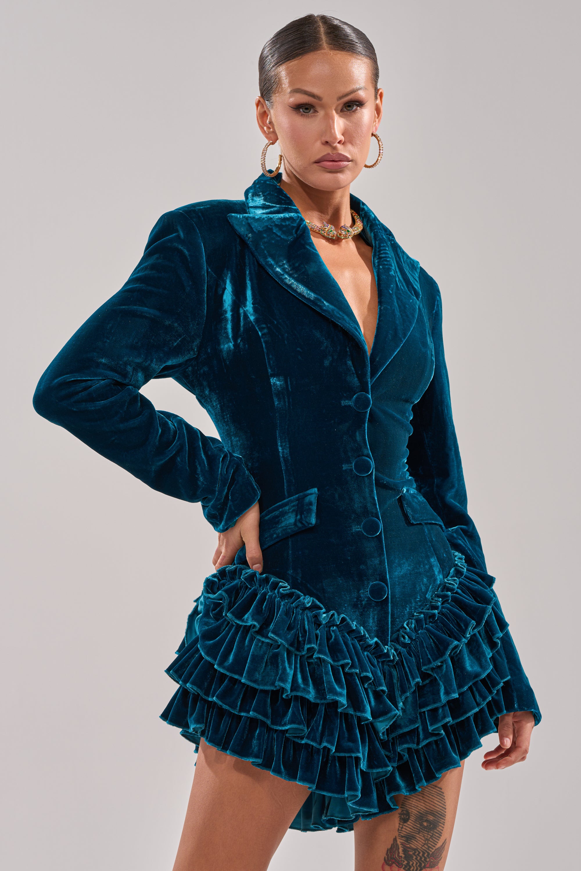 A woman wearing the SHE'S ROYALTY VELVET ROMPER in teal stands confidently against a neutral background, accessorized with slicked-back hair, gold hoop earrings, and a chunky gold necklace.