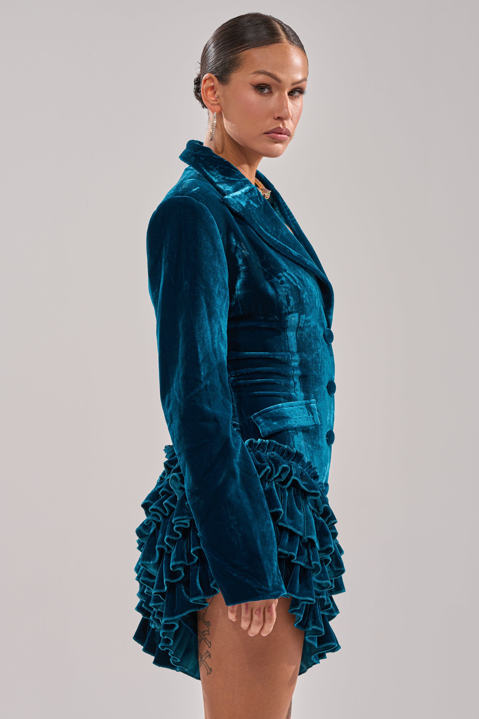 A woman in profile wears the SHE'S ROYALTY VELVET ROMPER in teal crushed velvet, featuring a layered ruffle hem. Her hair is slicked back, revealing earrings and a thigh tattoo, with a plain gray background behind her.