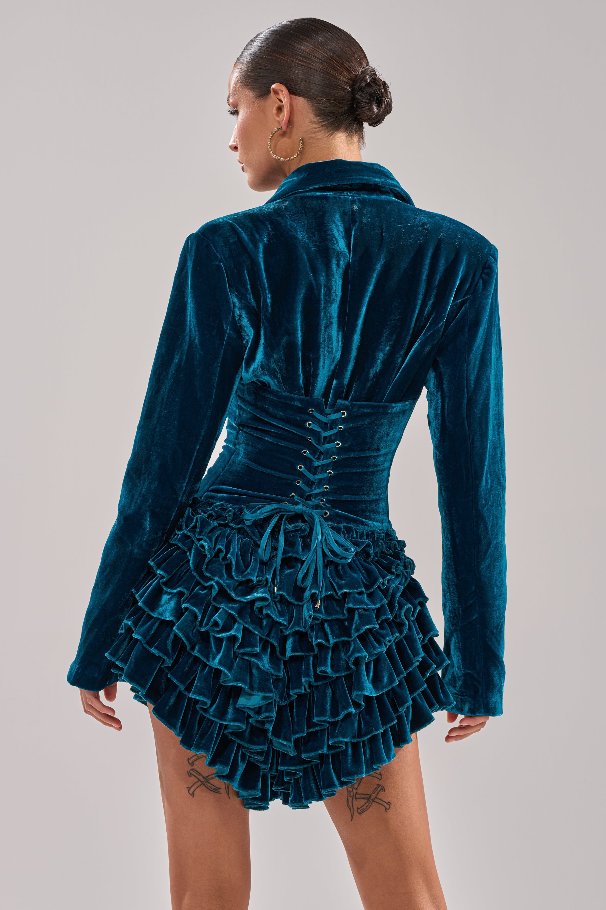 A woman with a low bun wears the SHE'S ROYALTY VELVET ROMPER in teal crushed velvet, featuring a fitted corset-style bodice, long sleeves, and a short layered ruffle hem. From the back, lace-up details and two thigh tattoos are visible.