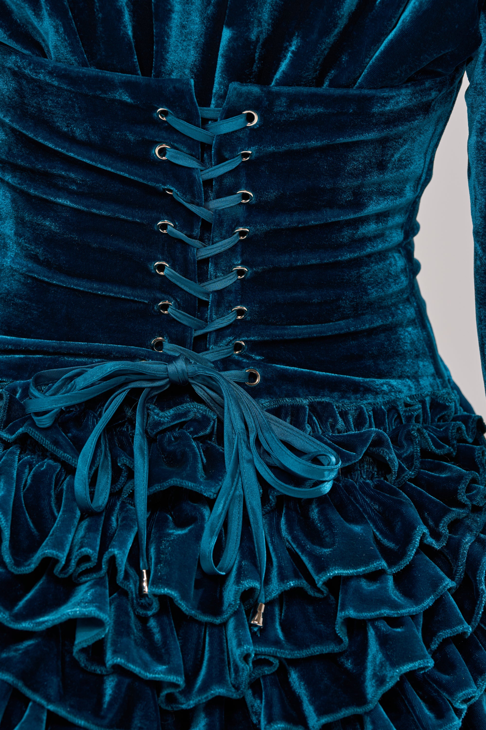 Close-up of the SHE'S ROYALTY VELVET ROMPER in teal crushed velvet, featuring a corset-style lace-up bodice with silver eyelets and a bow above a ruffled, layered skirt with a delicate ruffle hemline.