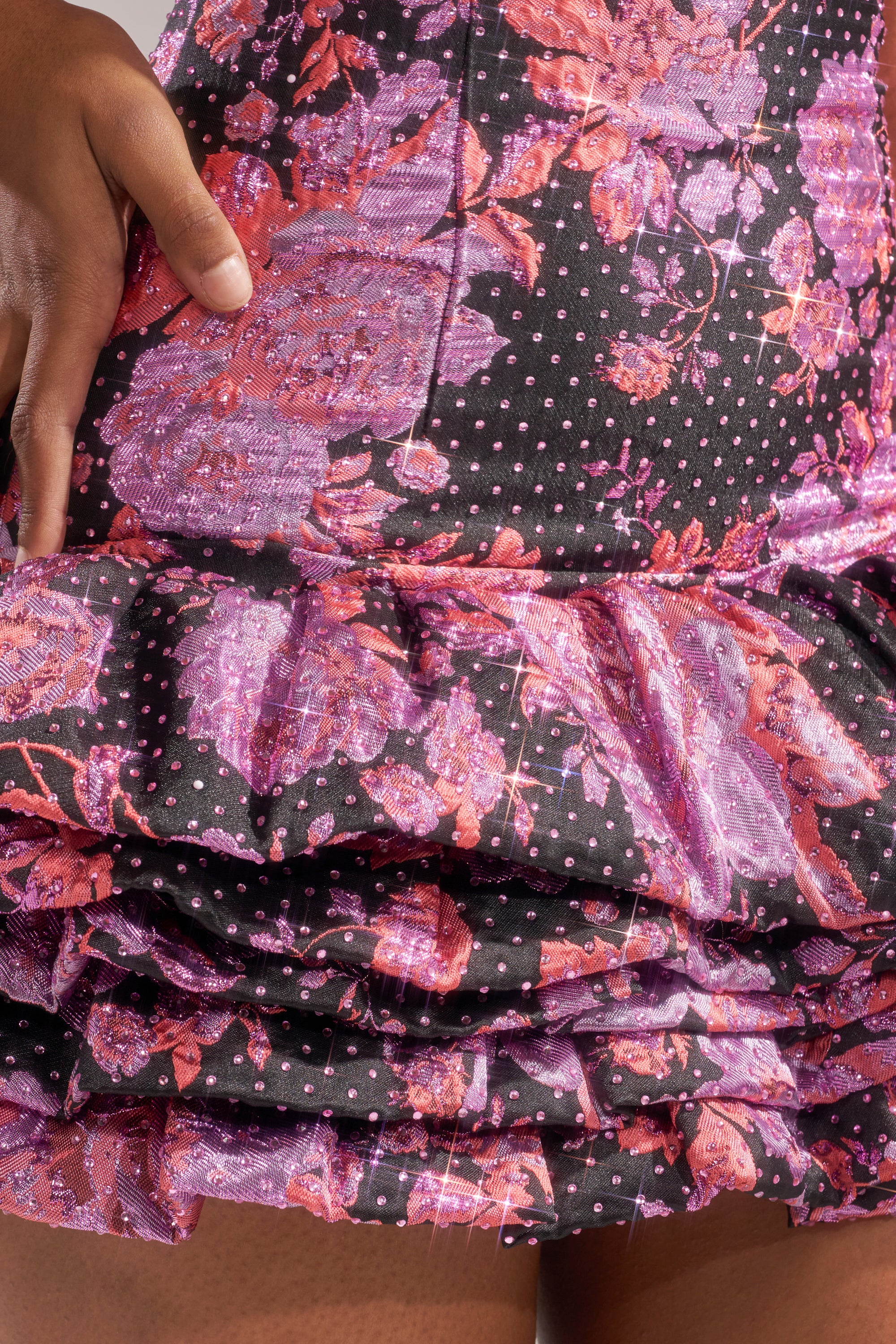 A close-up of a hand resting on the ruffled skirt of the GIRLS WHO BRUNCH BROCADE MINI DRESS, featuring a pink and purple floral jacquard pattern on a black background with small white spots. 8