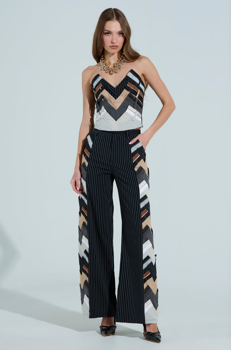 A woman exudes office chic vibes in a chevron-patterned top and STANDING ON BUSINESS LAYERED TROUSERS featuring pinstriped wide-leg design with matching chevron panels. She completes her stylish look with a chunky necklace and black heels.
