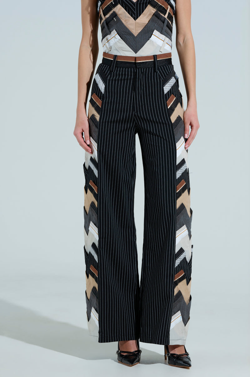 A person exudes office chic in a geometric-patterned top and STANDING ON BUSINESS LAYERED TROUSER with high-waisted, wide-leg stripes and brown, beige, black angular details. They stand confidently against a plain light background, embodying style and business-casual flair.
