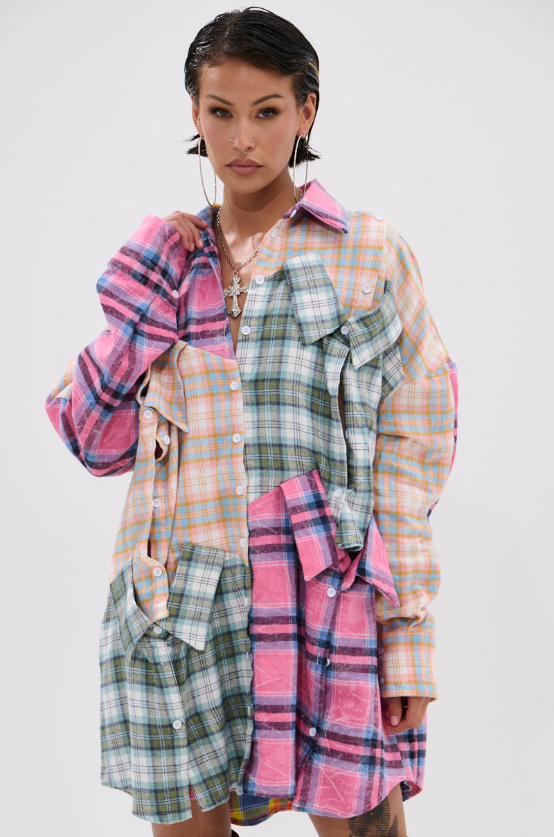 A person with short dark hair models the WILD CHILD PLAID SHIRT DRESS, an oversized long-sleeve patchwork shirt in pink, green, yellow, and blue plaids, standing against a plain white background.