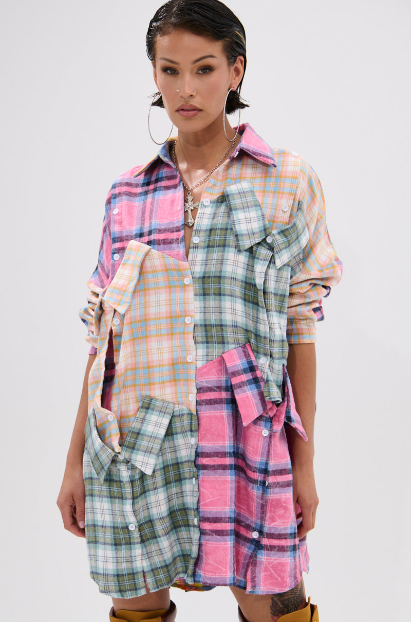 A woman wears the WILD CHILD PLAID SHIRT DRESS, an oversized fit with vibrant pink, green, blue, and yellow patchwork, extra sleeves and pockets on the front. She pairs it with slicked-back hair and large hoop earrings.