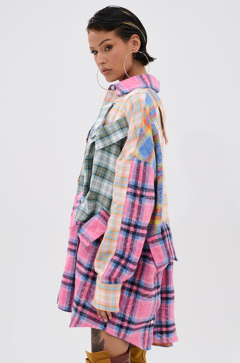 A woman with slicked-back hair and hoop earrings models the WILD CHILD PLAID SHIRT DRESS—an oversized patchwork plaid shirt in pastel pink, green, yellow, and blue—while looking over her shoulder against a white background.