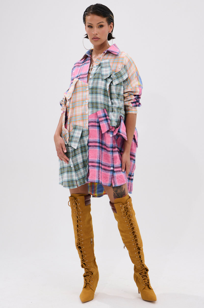 A woman stands confidently in the oversized WILD CHILD PLAID SHIRT DRESS with patchwork plaid, large pockets, and long sleeves, styled with tall mustard yellow lace-up boots. Her hair has a wet-look finish and she wears a neutral expression.