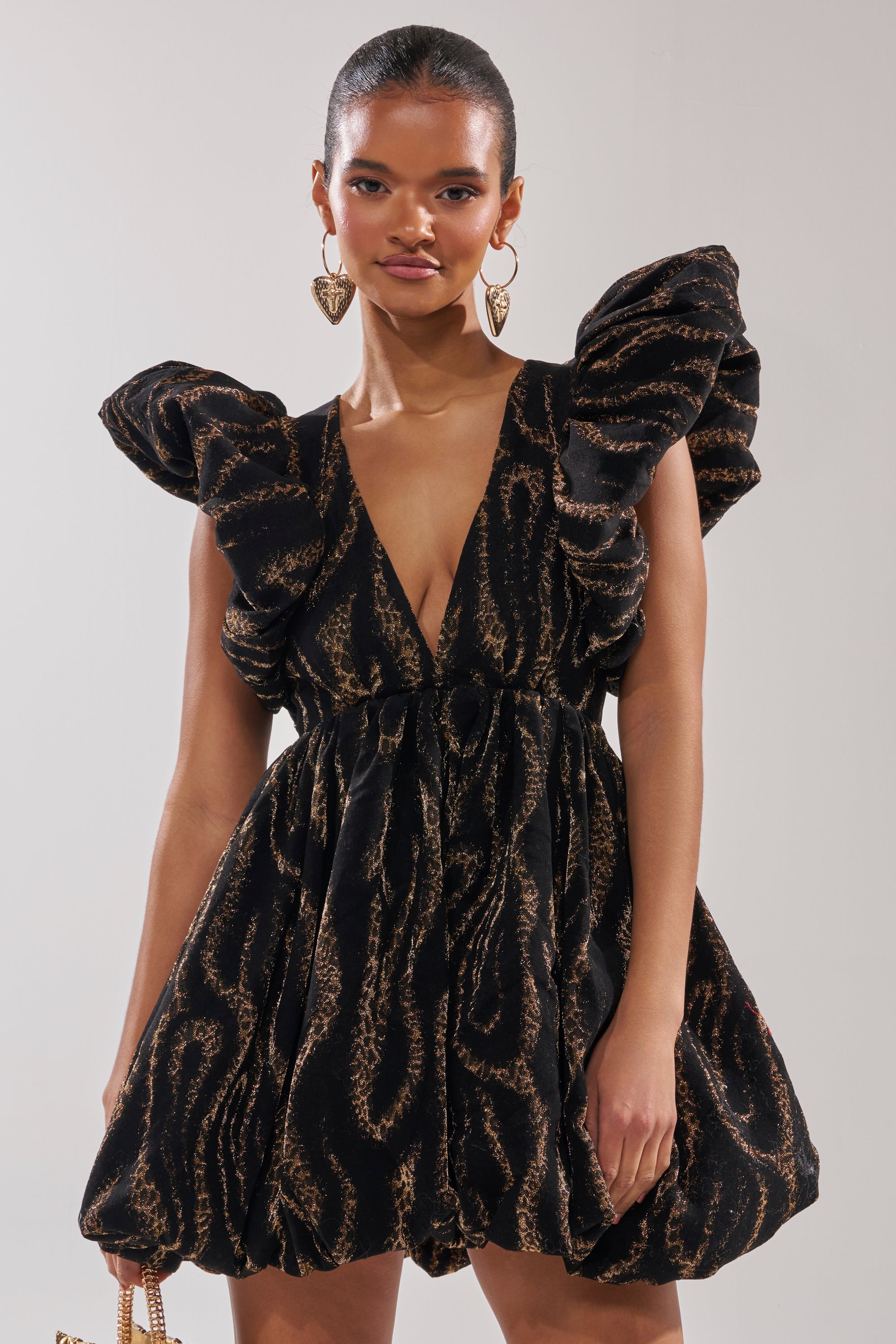 A woman wears the WILD HEART BROCADE BUBBLE MINI DRESS, featuring black and gold brocade, puffed sleeves, a deep V-neckline, and a flared skirt—ideal for holiday parties. She pairs it with large heart-shaped earrings and a small bag.