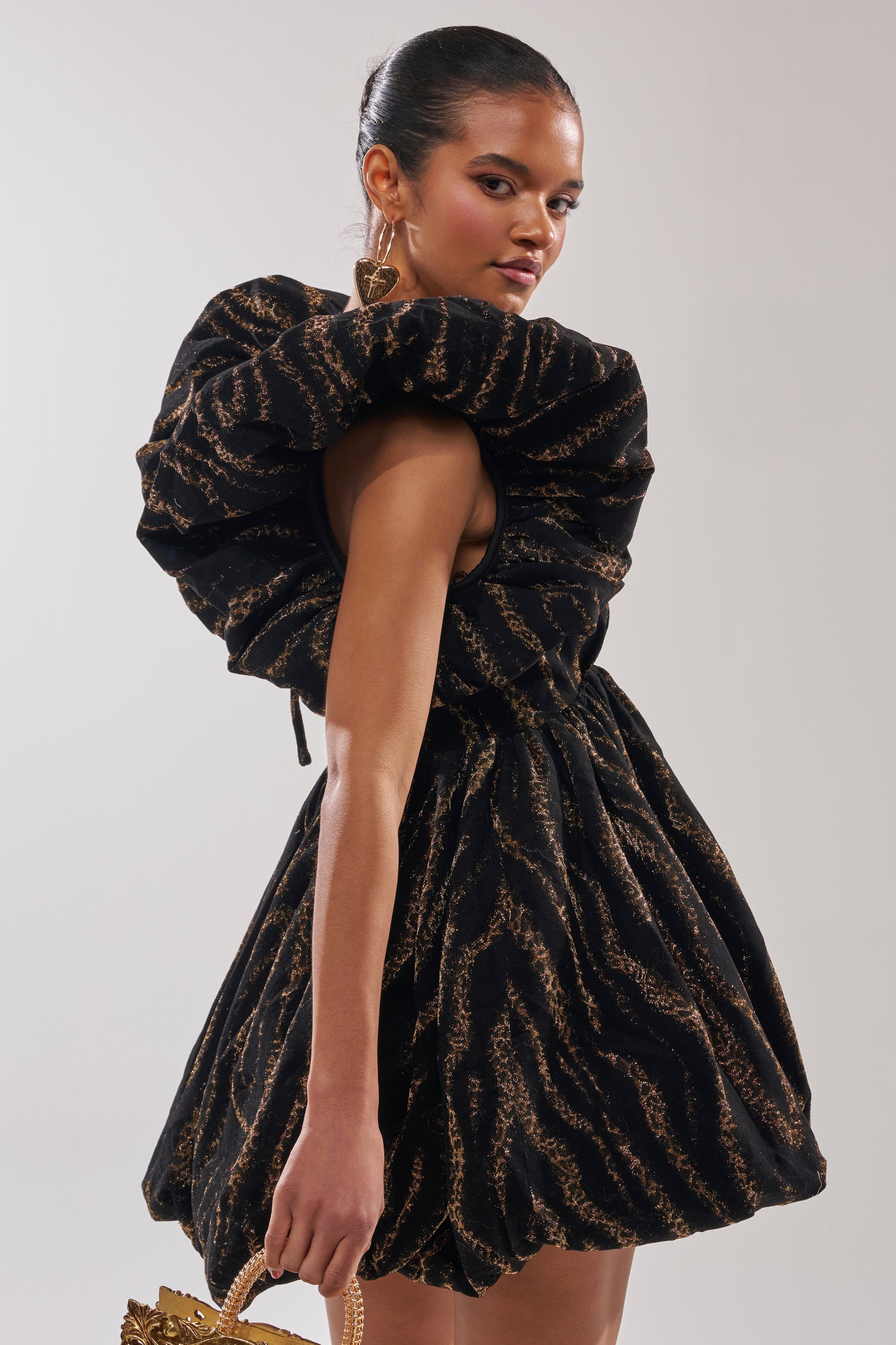 A woman stands sideways in the WILD HEART BROCADE BUBBLE MINI DRESS, featuring dramatic ruffled sleeves and a voluminous skirt. Her hair is tied back; she accessorizes with a gold handbag and statement gold earrings.