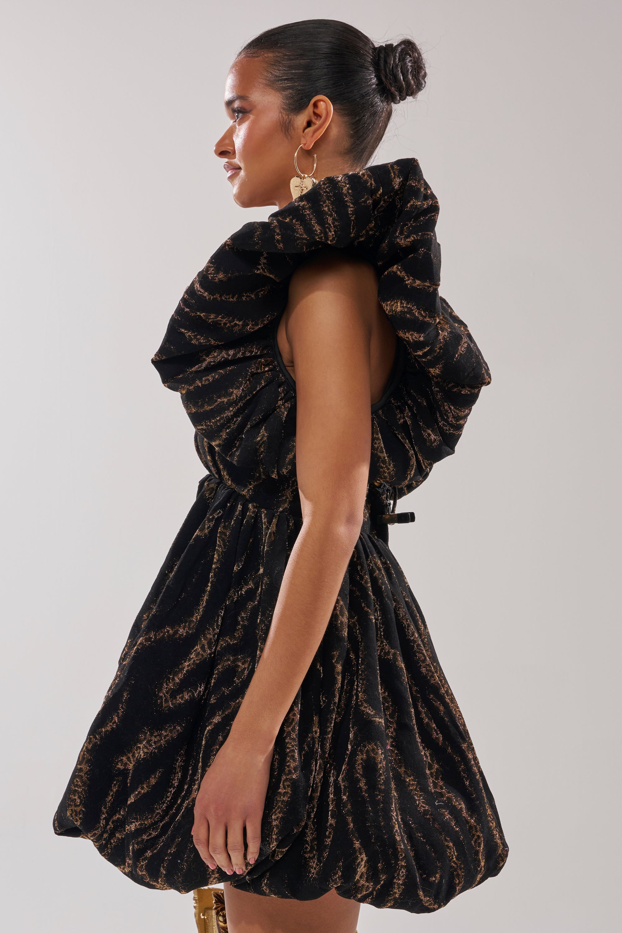 A woman stands in profile wearing the WILD HEART BROCADE BUBBLE MINI DRESS, a gold metallic mini with a ruffled collar and bubble hem—ideal for holiday parties. Her hair is styled in a sleek bun with large round earrings.