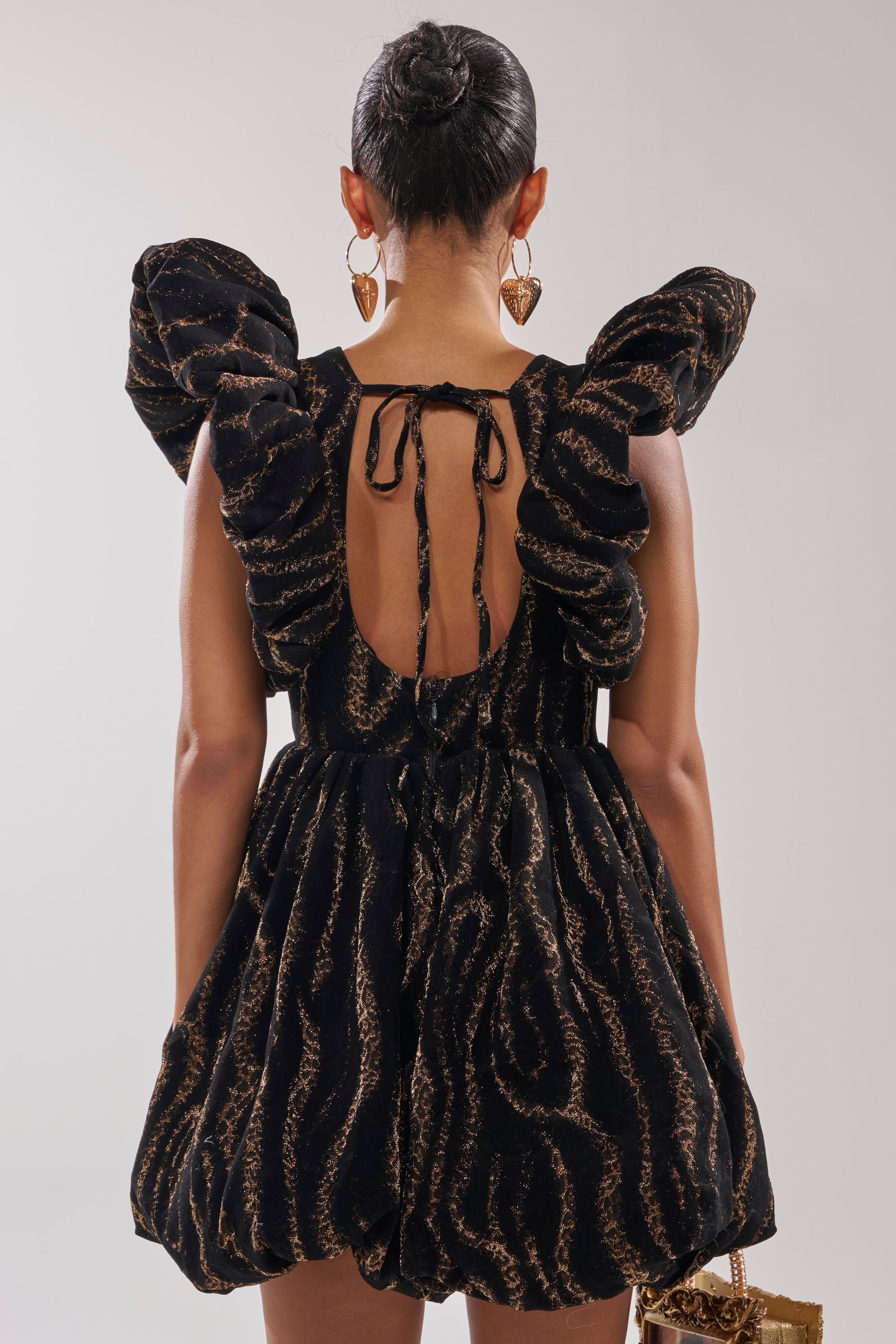 A woman is seen from behind in the WILD HEART BROCADE BUBBLE MINI DRESS with gold patterns, carrying a gold bag, wearing large heart-shaped earrings, and sporting a sleek bun—an ideal holiday party look.