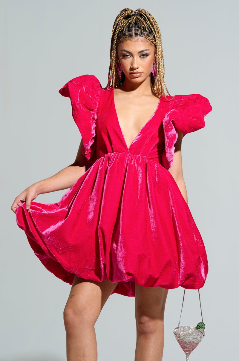 Wearing the BELLE OF THE BALL VELVET MINI DRESS in a vibrant pink hue, a person poses confidently. The dress features puffed shoulder detailing and a deep V-neckline. With long braided hair and holding a small, ornate bag, they stand against a minimalist gray background.