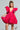 Wearing the BELLE OF THE BALL VELVET MINI DRESS in a vibrant pink hue, a person poses confidently. The dress features puffed shoulder detailing and a deep V-neckline. With long braided hair and holding a small, ornate bag, they stand against a minimalist gray background.