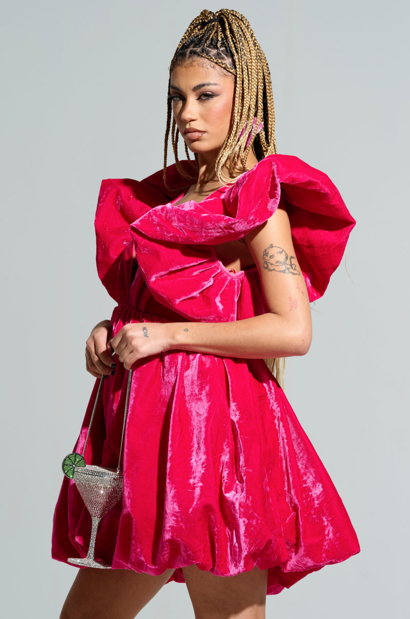 A person with braided hair poses confidently in the BELLE OF THE BALL VELVET MINI DRESS, a vibrant red outfit featuring puffed shoulder detailing. They are holding a sparkling cocktail glass with a green stem, while their tattoos are visible on their left arm against a plain background.