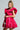 A person with braided hair poses confidently in the BELLE OF THE BALL VELVET MINI DRESS, a vibrant red outfit featuring puffed shoulder detailing. They are holding a sparkling cocktail glass with a green stem, while their tattoos are visible on their left arm against a plain background.