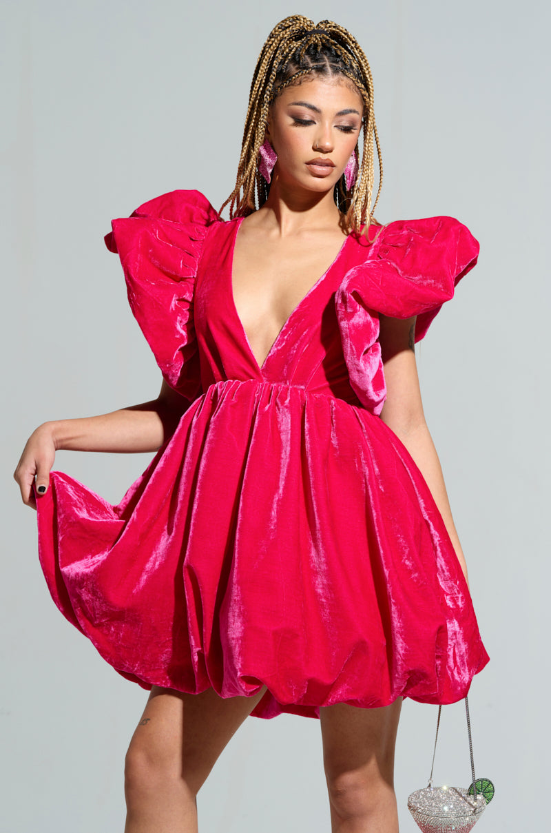 Dressed in the stunning BELLE OF THE BALL VELVET MINI DRESS, a woman gracefully poses, showcasing its vibrant pink hue with large ruffled sleeves and a deep V-neckline featuring puffed shoulder detailing. She holds the hem of her dress in one hand while clutching a heart-shaped purse in the other, her braided hair perfectly complementing her elegant ensemble.