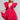 Dressed in the stunning BELLE OF THE BALL VELVET MINI DRESS, a woman gracefully poses, showcasing its vibrant pink hue with large ruffled sleeves and a deep V-neckline featuring puffed shoulder detailing. She holds the hem of her dress in one hand while clutching a heart-shaped purse in the other, her braided hair perfectly complementing her elegant ensemble.