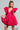 Dressed in the stunning BELLE OF THE BALL VELVET MINI DRESS, a woman gracefully poses, showcasing its vibrant pink hue with large ruffled sleeves and a deep V-neckline featuring puffed shoulder detailing. She holds the hem of her dress in one hand while clutching a heart-shaped purse in the other, her braided hair perfectly complementing her elegant ensemble.