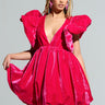 Dressed in the stunning BELLE OF THE BALL VELVET MINI DRESS, a woman gracefully poses, showcasing its vibrant pink hue with large ruffled sleeves and a deep V-neckline featuring puffed shoulder detailing. She holds the hem of her dress in one hand while clutching a heart-shaped purse in the other, her braided hair perfectly complementing her elegant ensemble.