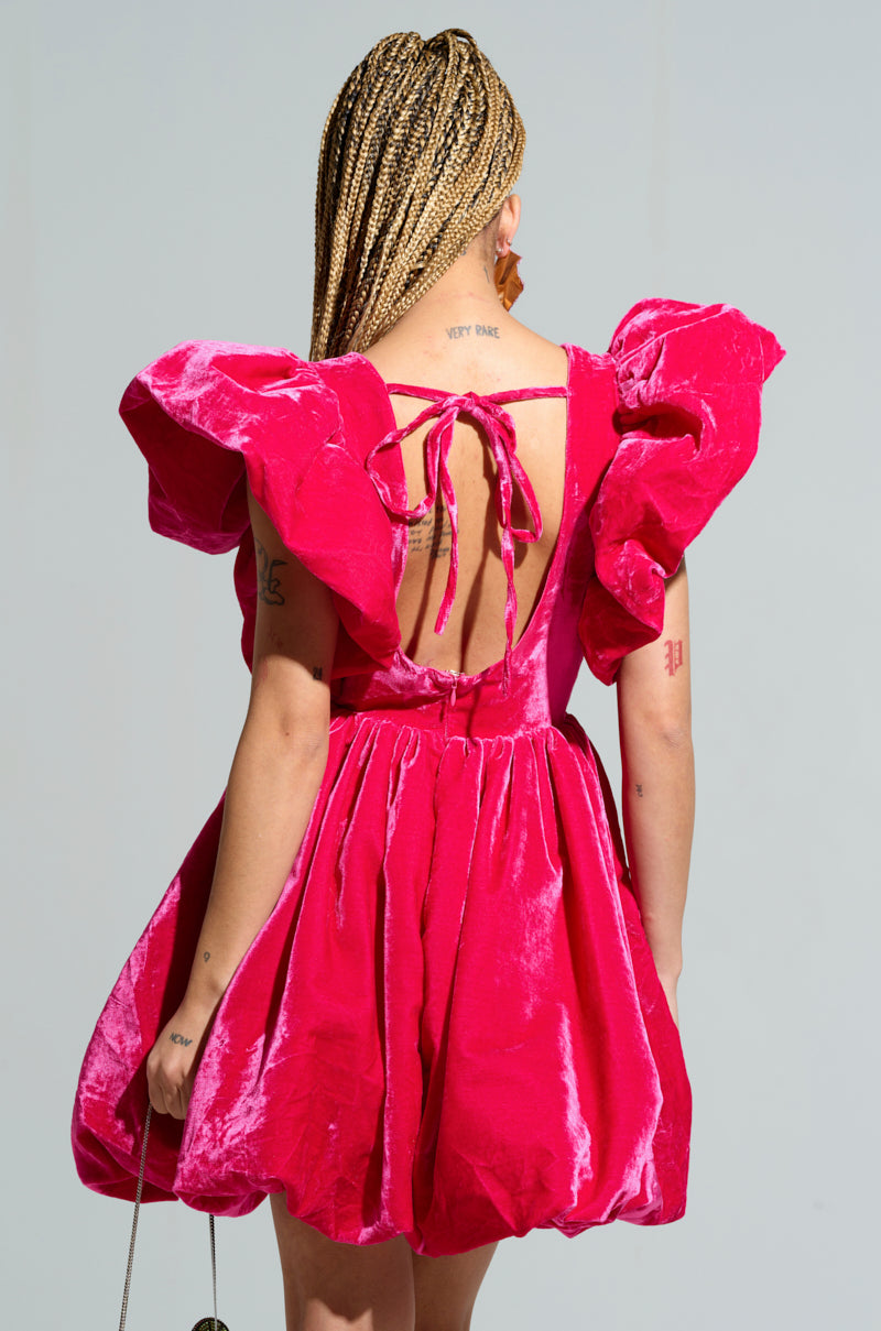 With braided hair, a person confidently showcases the BELLE OF THE BALL VELVET MINI DRESS, which features a vibrant pink color, puffed shoulder details, and an open back, standing against a simple background.