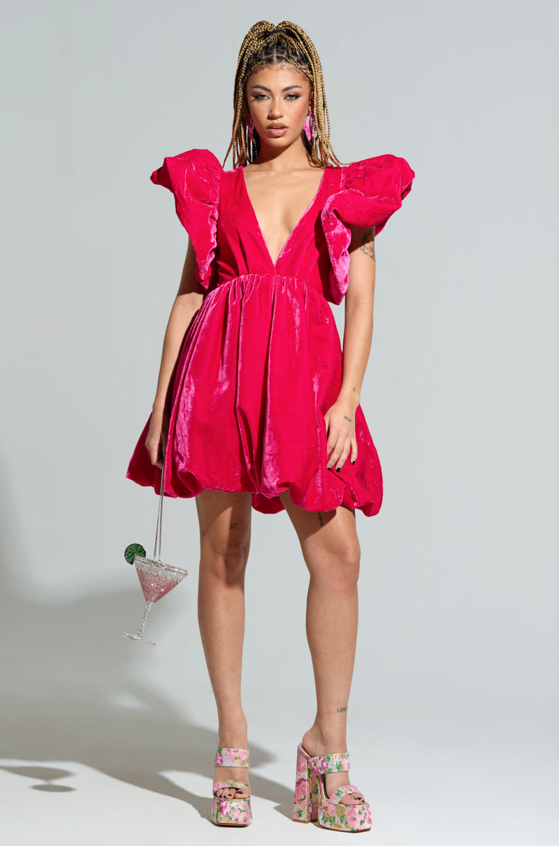 A person with braided hair is wearing the BELLE OF THE BALL VELVET MINI DRESS in bright pink, showcasing puffed shoulder detailing. They are also wearing floral platform heels and holding a cocktail glass with a lime garnish against a plain gray background.