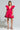 A person with braided hair is wearing the BELLE OF THE BALL VELVET MINI DRESS in bright pink, showcasing puffed shoulder detailing. They are also wearing floral platform heels and holding a cocktail glass with a lime garnish against a plain gray background.
