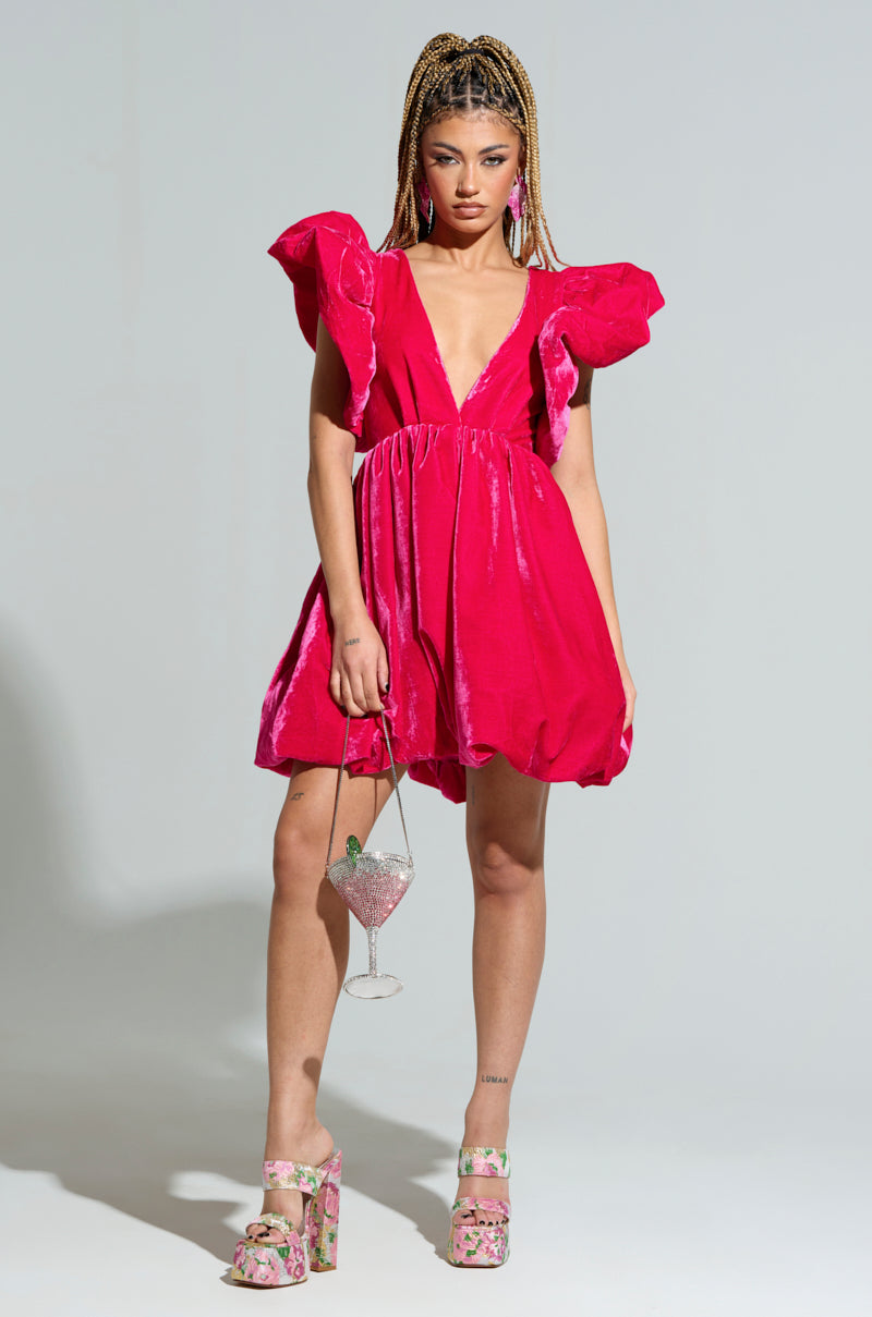 A woman poses confidently in the BELLE OF THE BALL VELVET MINI DRESS, showcasing its bright pink hue, bold ruffled sleeves, and deep V-neck. She accessorizes with a sparkly, transparent handbag shaped like a drink glass and floral platform shoes. Her hair is styled in braids, complementing the outfit's elegant puffed shoulder detailing.