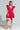 A woman poses confidently in the BELLE OF THE BALL VELVET MINI DRESS, showcasing its bright pink hue, bold ruffled sleeves, and deep V-neck. She accessorizes with a sparkly, transparent handbag shaped like a drink glass and floral platform shoes. Her hair is styled in braids, complementing the outfit's elegant puffed shoulder detailing.